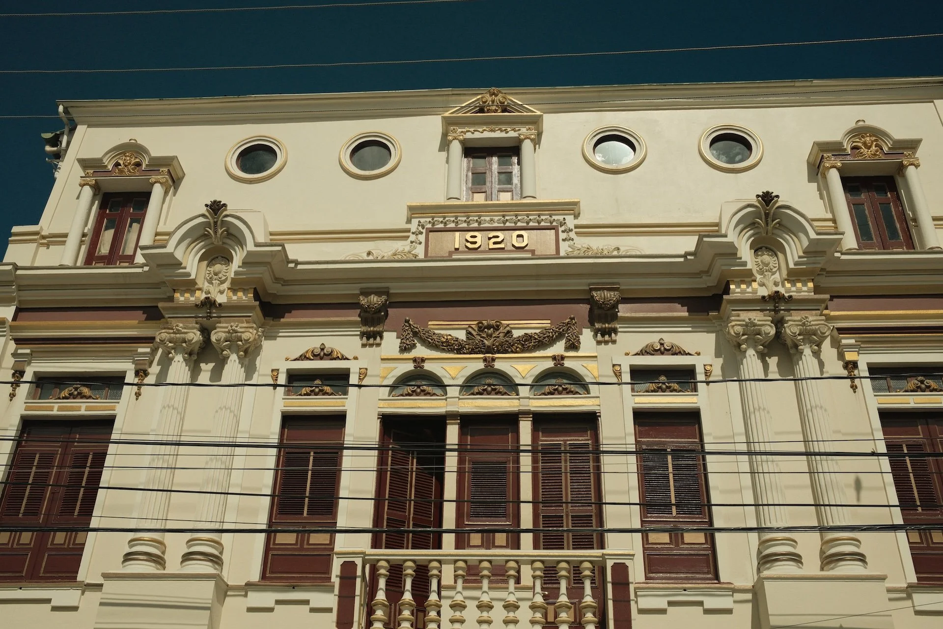 Historic building with intricate architectural details, the year 1920 displayed on a plaque, multiple windows with closed shutters, and decorative elements such as garlands and ornate columns.