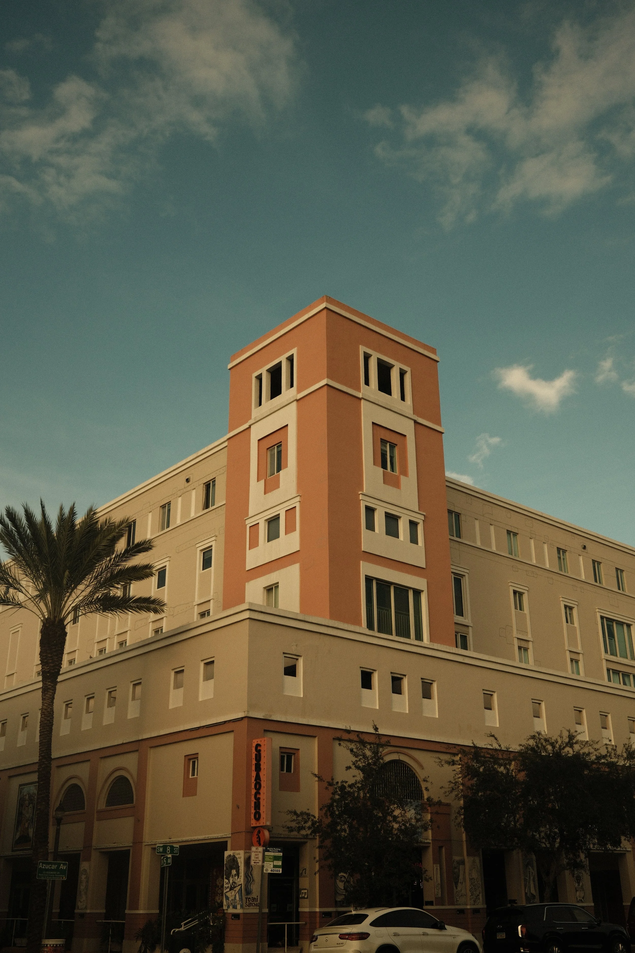 A multi-story building with pastel pink and beige colors, featuring a tower-like structure at the corner, with the sign 'GUITAR CENTER' near the entrance, palm trees, parked cars, and a clear sky with scattered clouds.