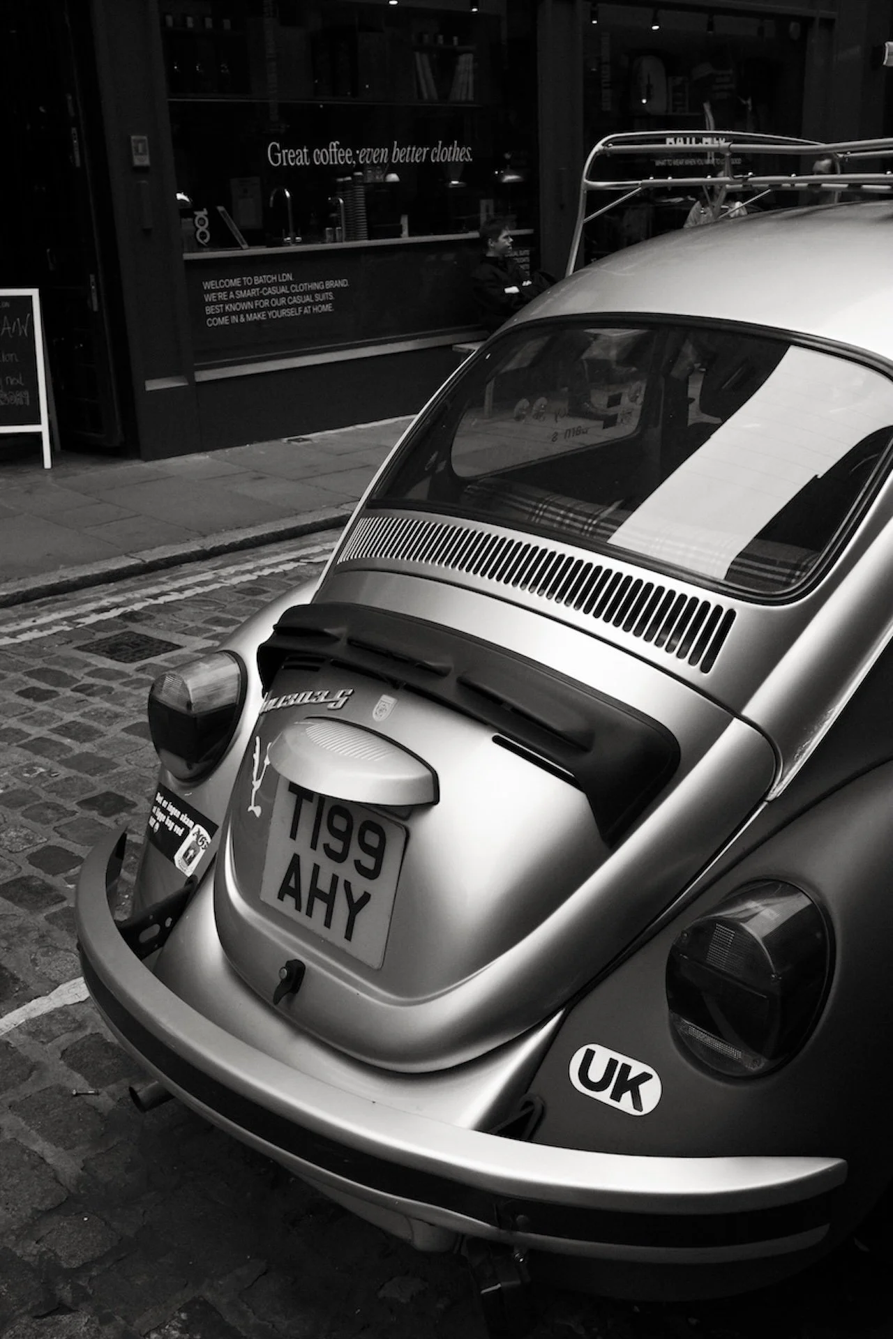A classic silver Volkswagen Beetle parked on a cobblestone street in front of a café. The car has UK license plates, a sticker that reads 'UK', and is reflected in the café window.