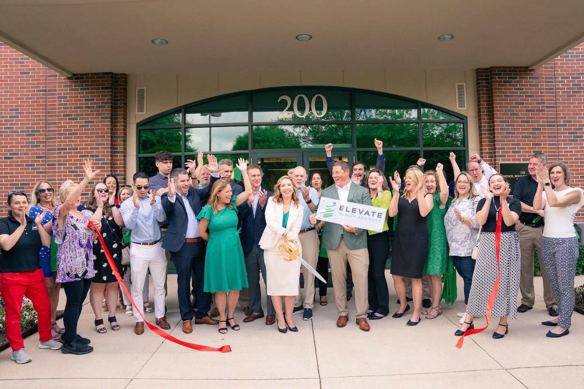 Group of people celebrating outside a building with the number 200 on the front, some holding a ribbon and a large pair of scissors, indicating a ribbon-cutting event.