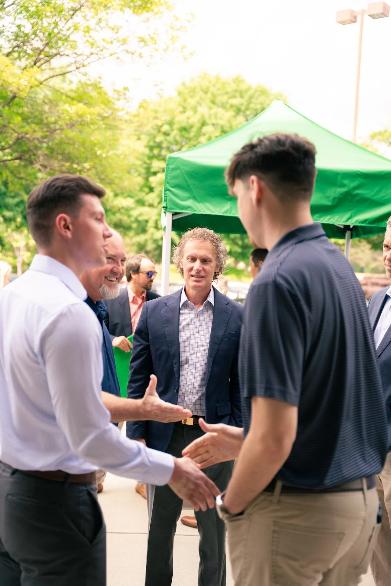 Group of men in business attire engaged in conversation at an outdoor event with green trees and a green canopy in the background.
