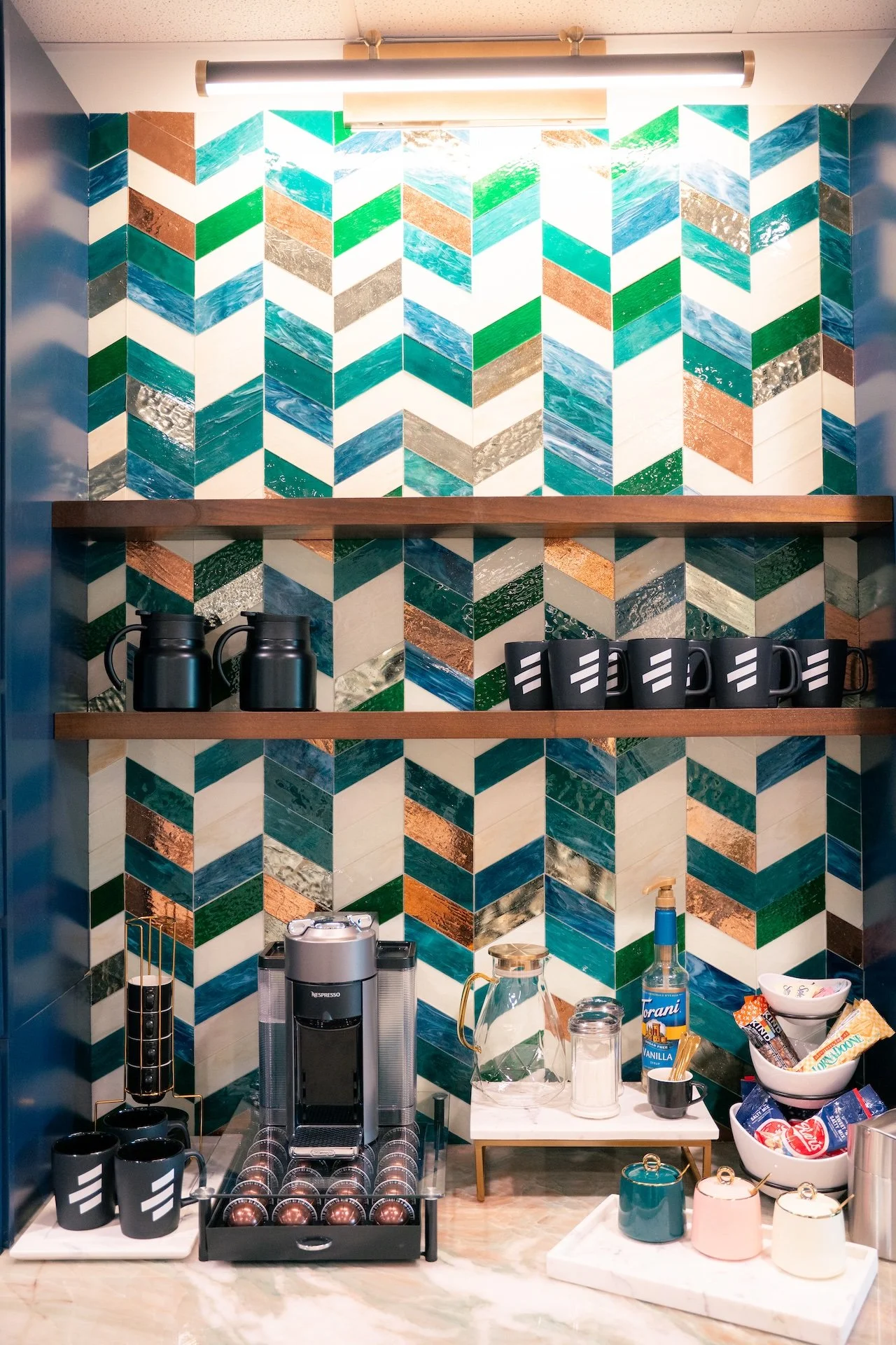 Coffee station with a colorful geometric tile backsplash, a Nespresso coffee machine, black cups, mugs, bowls with snacks, and various coffee supplies.