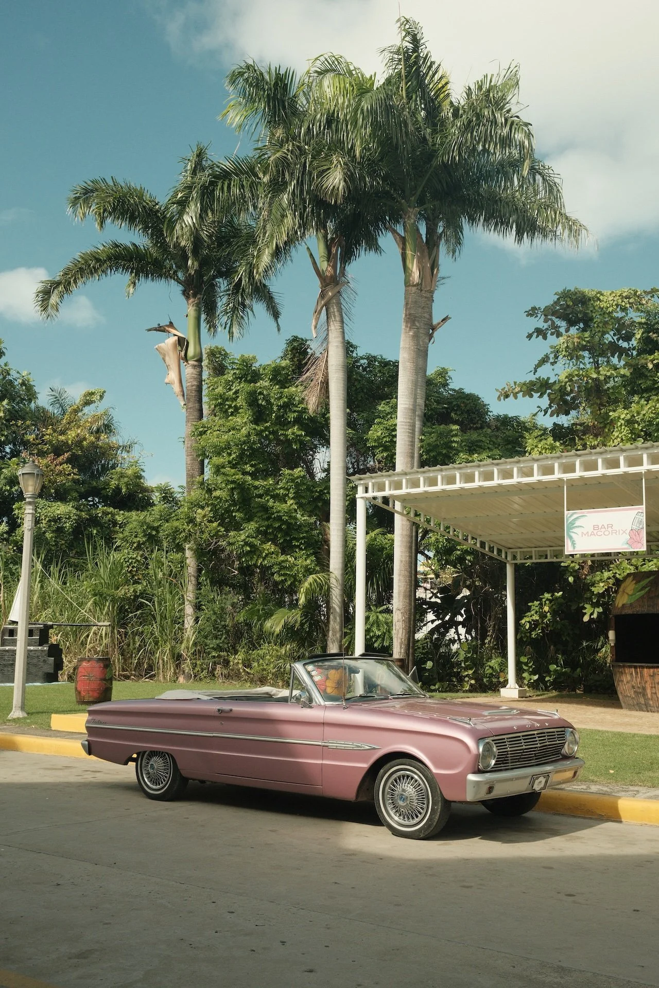 A vintage pink convertible car parked on a street near a grassy area with palm trees and lush green foliage. A sign for a bar, 'Bar Macorix', is visible in the background under a white canopy.