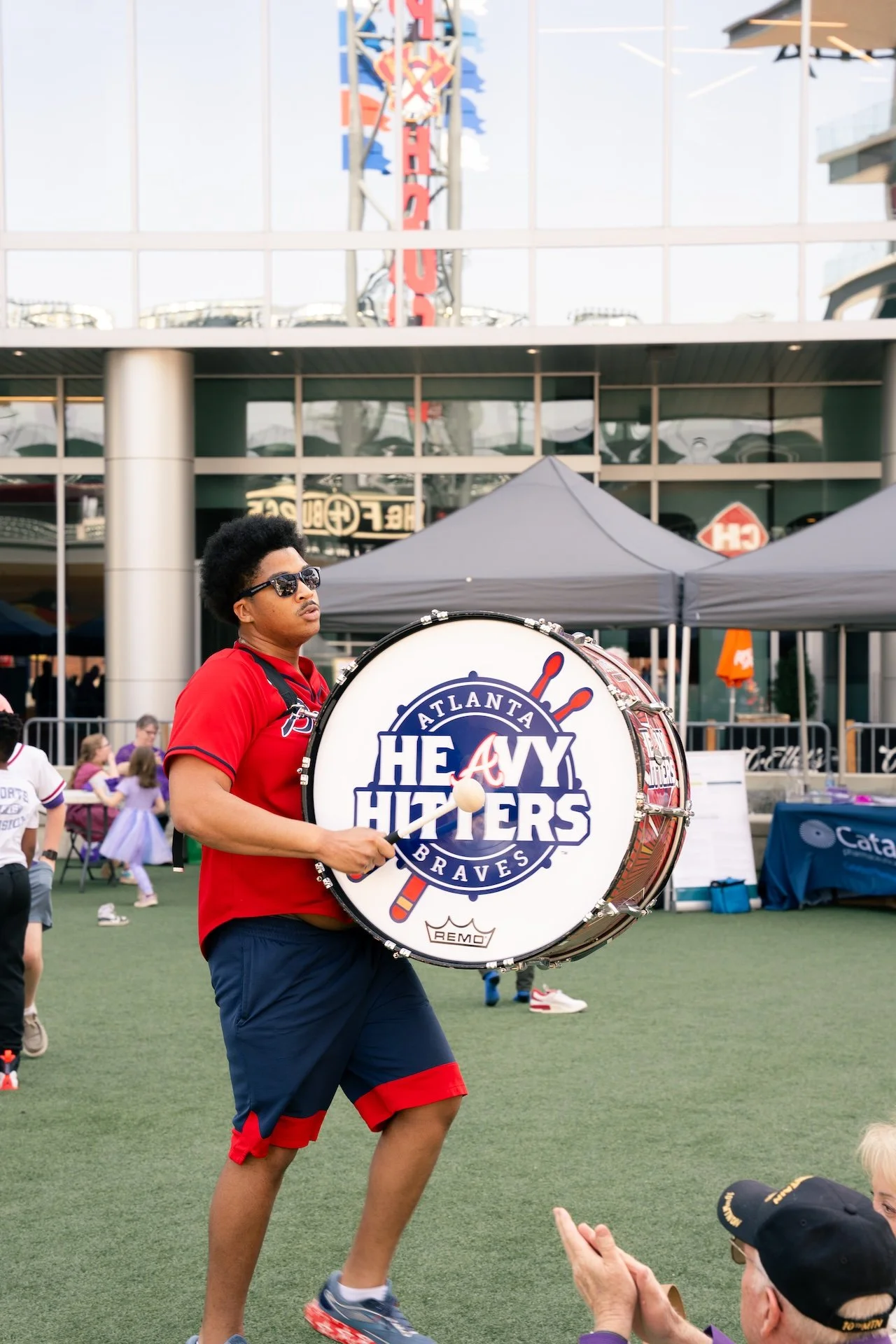 A man wearing sunglasses and a red Atlanta Heavy Hitters Braves shirt is playing a bass drum with the team's logo during a public event, with a background of tents, a building, and a few children.