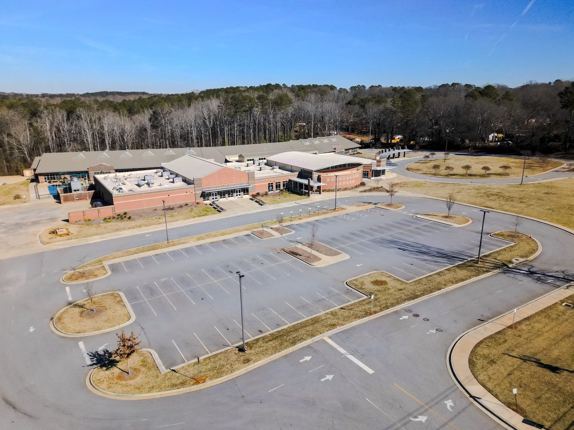 Empty parking lot in front of a large modern building with multiple sections, surrounded by trees and open grassy areas, under a clear blue sky.