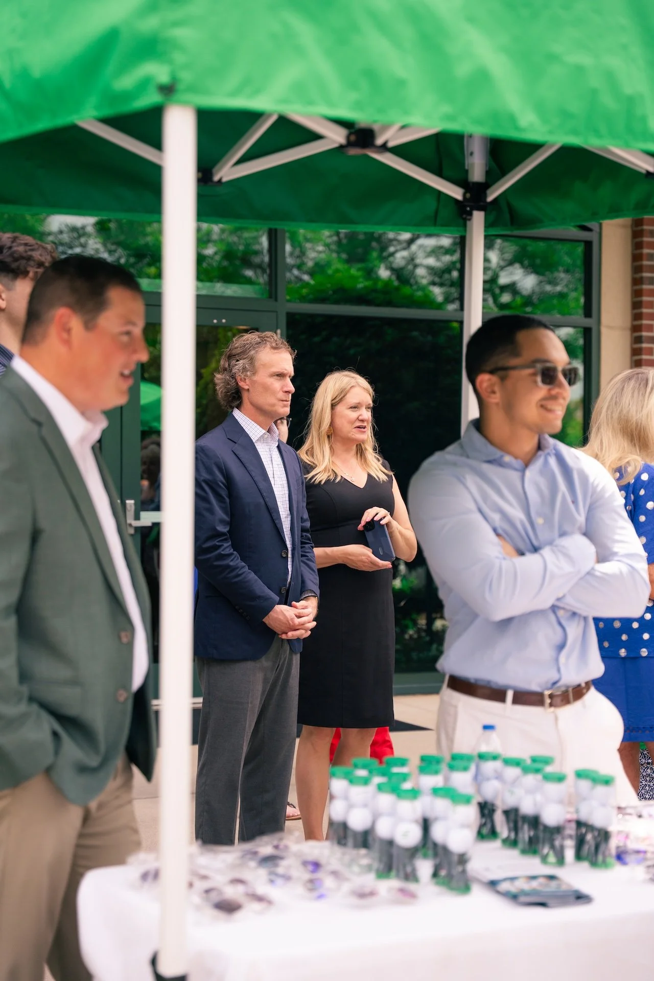 Group of professionally dressed people standing outdoors under a green canopy, with tables displaying water bottles and promotional items.