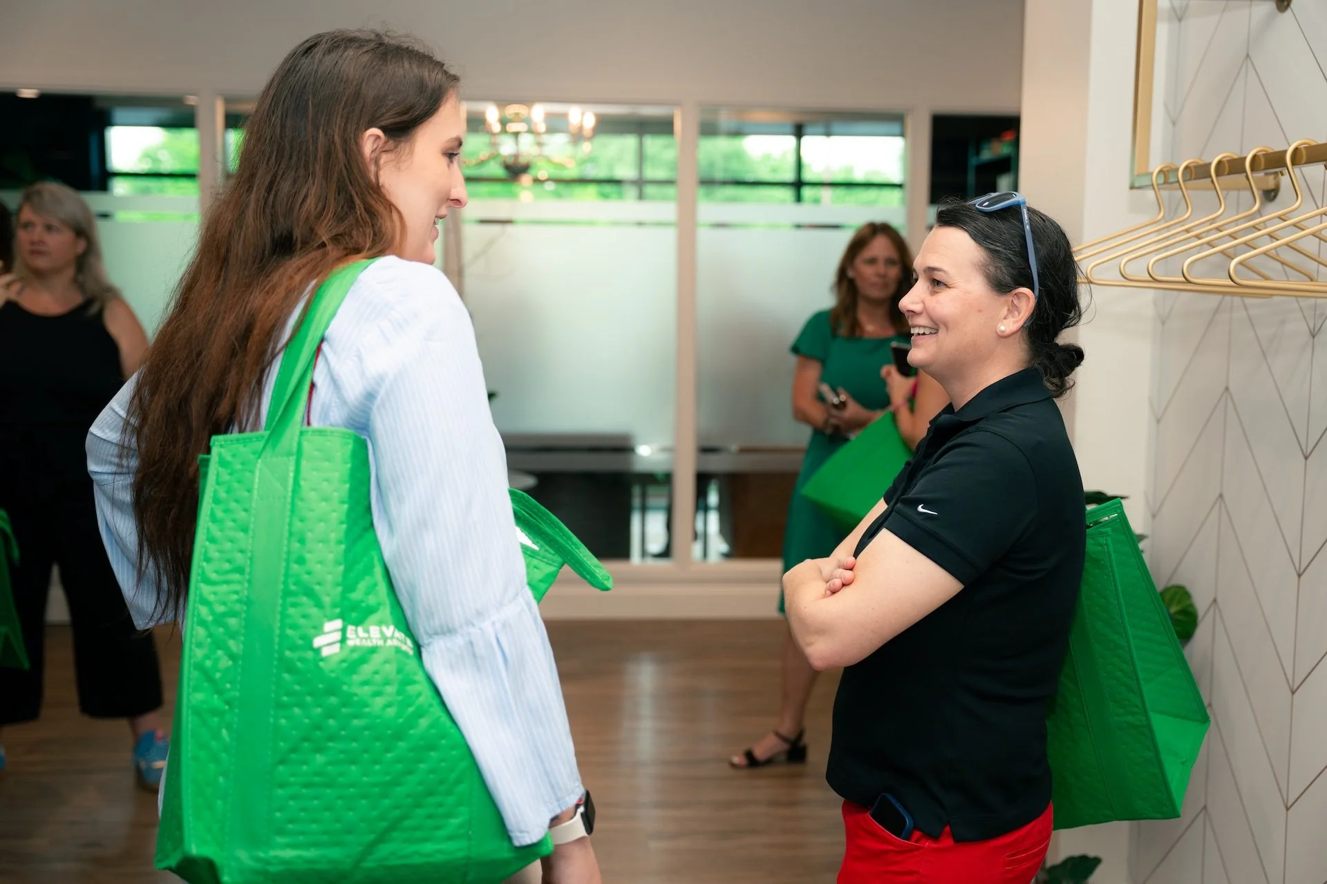 Two women, smiling and engaging in a conversation, standing inside a modern office or conference space, each carrying large green tote bags with the Elevar Health logo. In the background, other women are present, some holding green bags, and there is