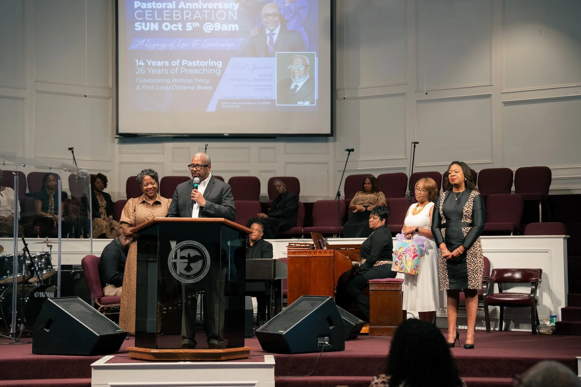 A pastor standing at a podium giving a speech during a church service, with a large screen behind him displaying details of a Pastoral Anniversary celebration on Sunday, October 5th at 9 am. Several women and men are seated or standing behind him, an
