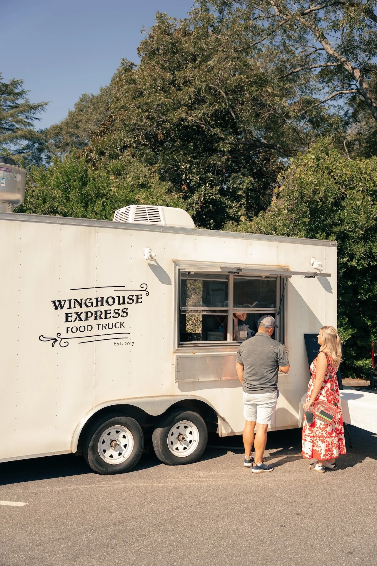 A white food truck named 'Winghouse Express Food Truck' with a window open, where an employee is serving customers. Two people are standing at the window, a man in a gray shirt and shorts, and a woman in a red floral dress with blonde hair, holding a