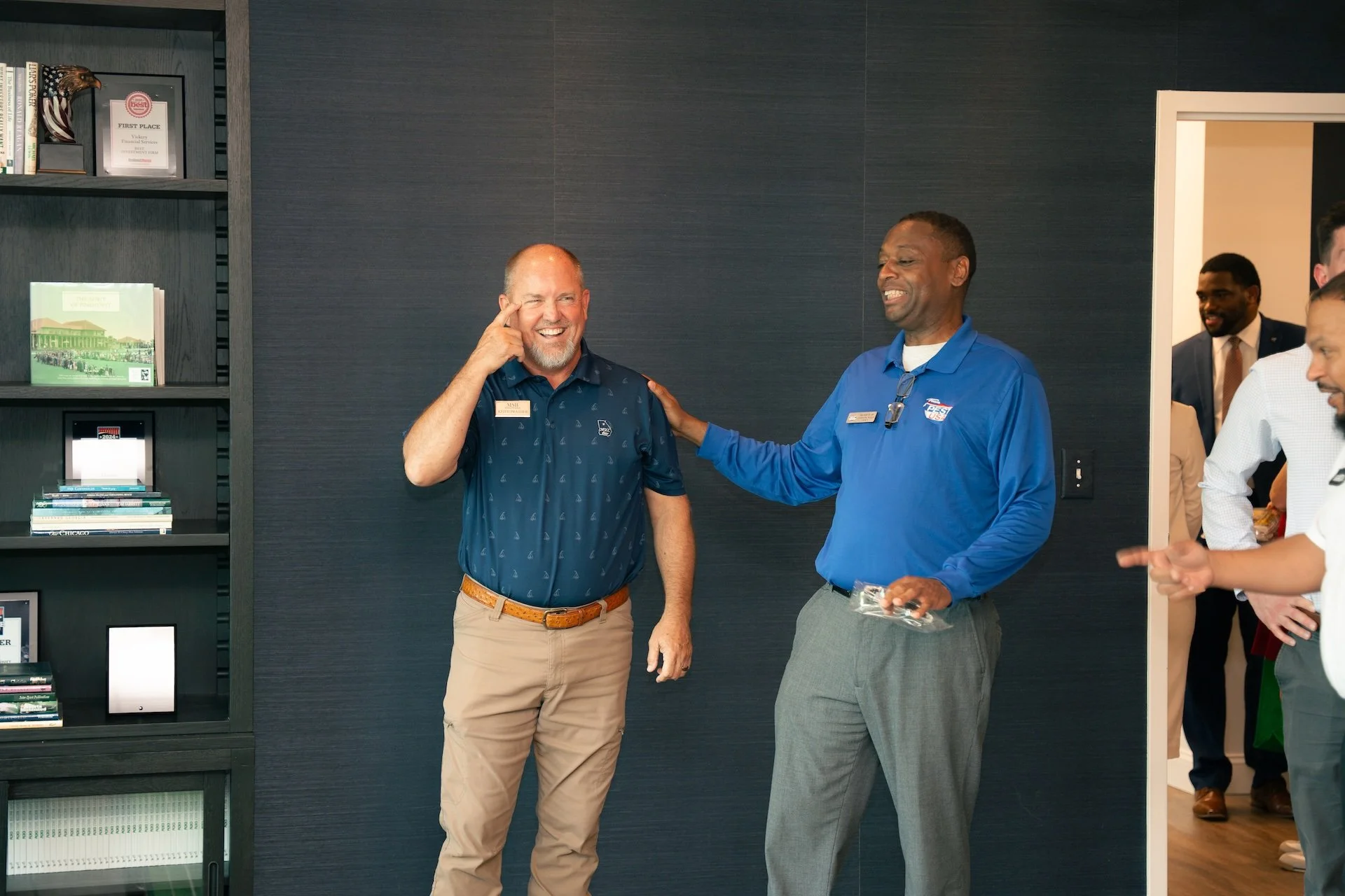 Two men smiling and talking in an office, one with his hand on the other's shoulder, and several others visible in the background.