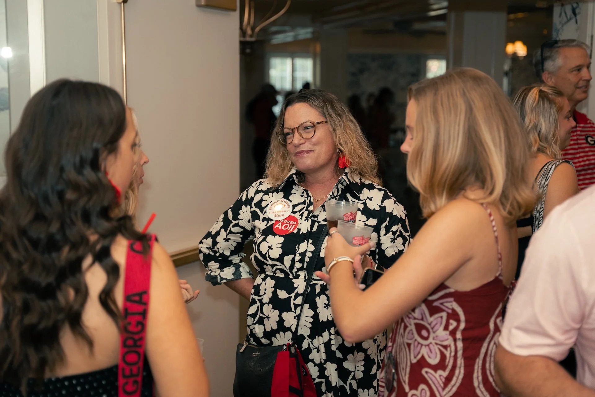Group of people socializing at an event, with women holding drinks and wearing casual summer attire, indoors in a well-lit room.