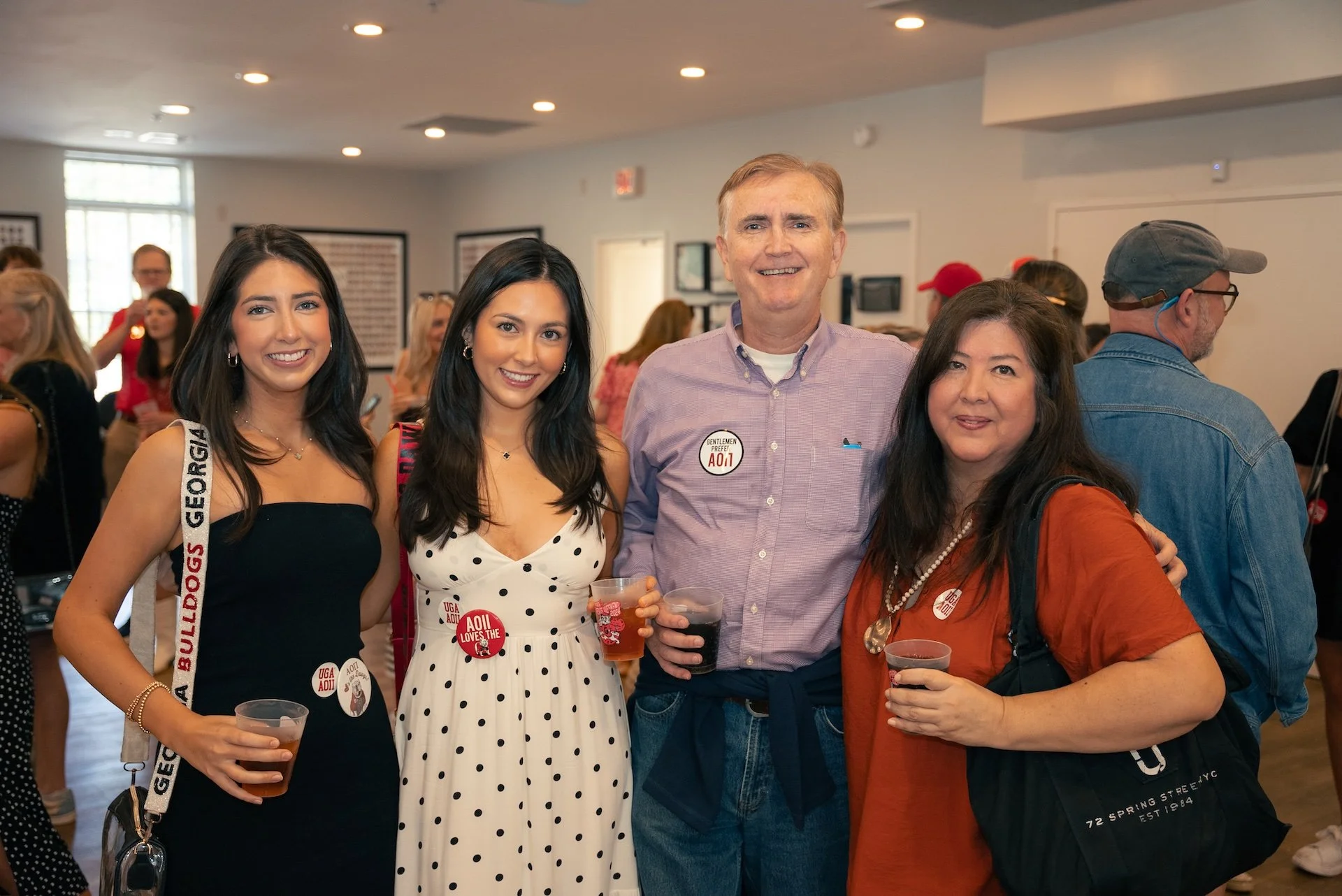 Group of four smiling people at a social event, holding drinks, with other attendees in the background.