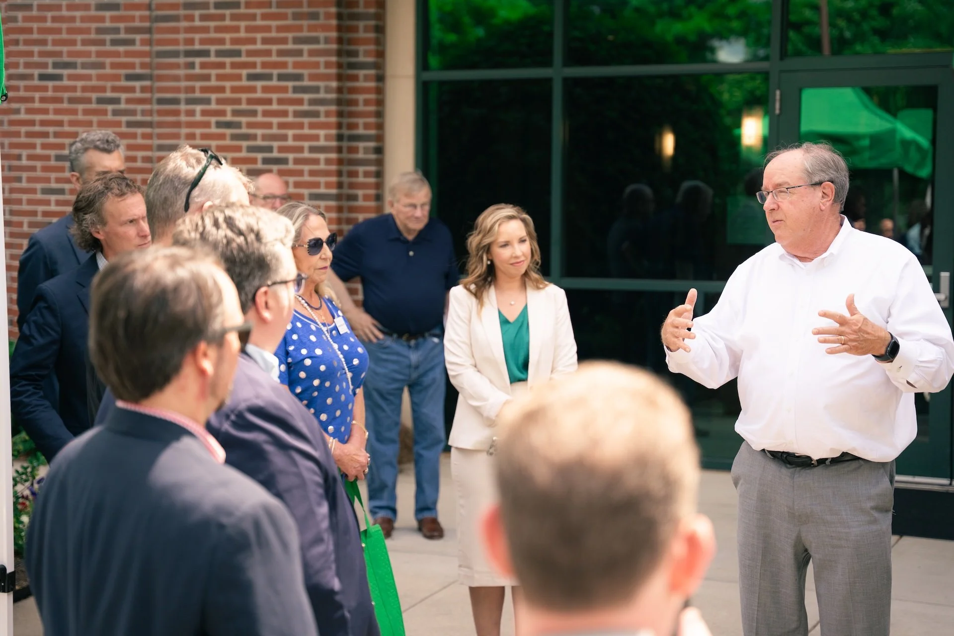 A man in white shirt and grey pants speaking to a group of people outdoors, with a brick wall and glass door behind them.