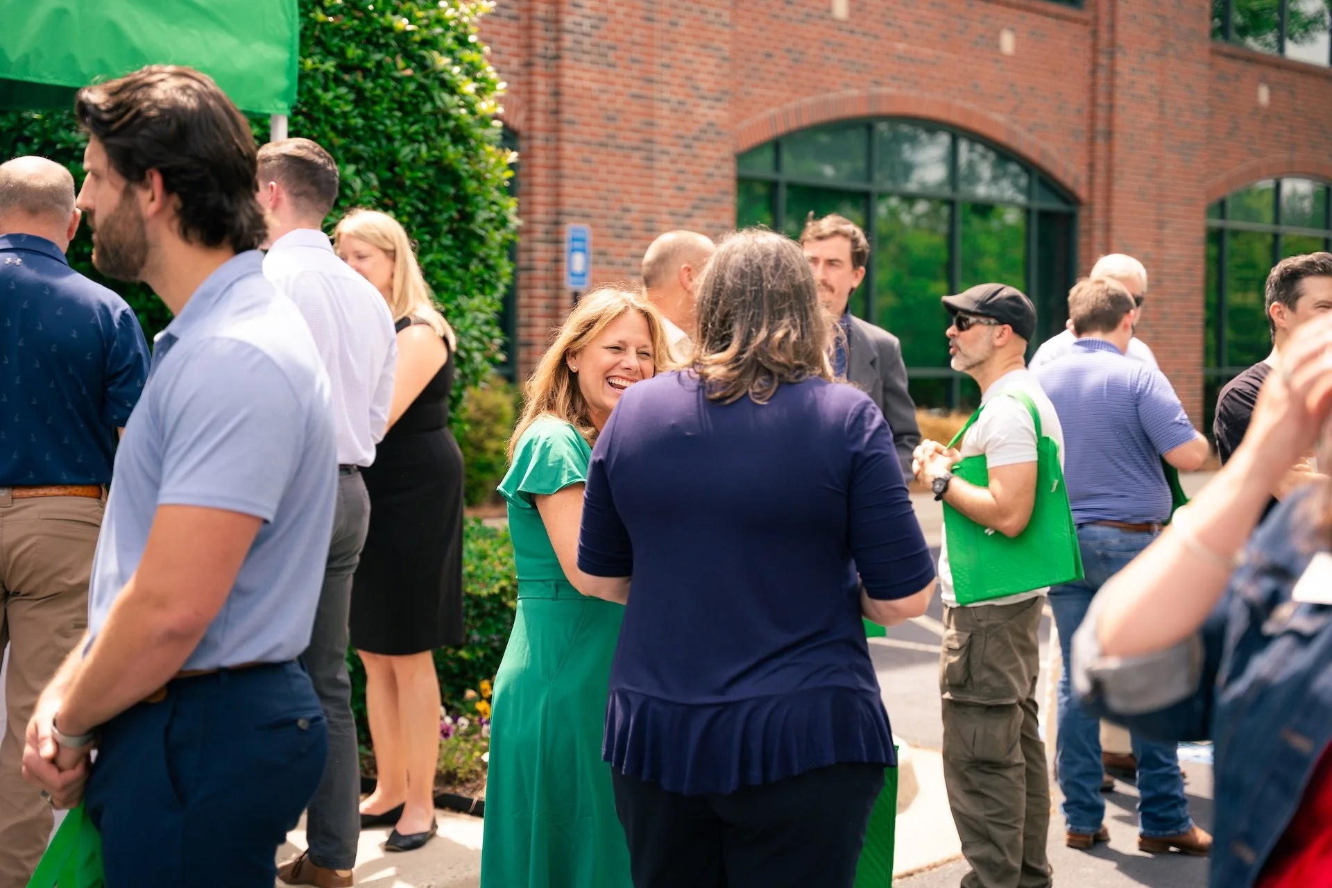 A group of people gathered outdoors in front of a brick building, engaging in conversations during a sunny day.