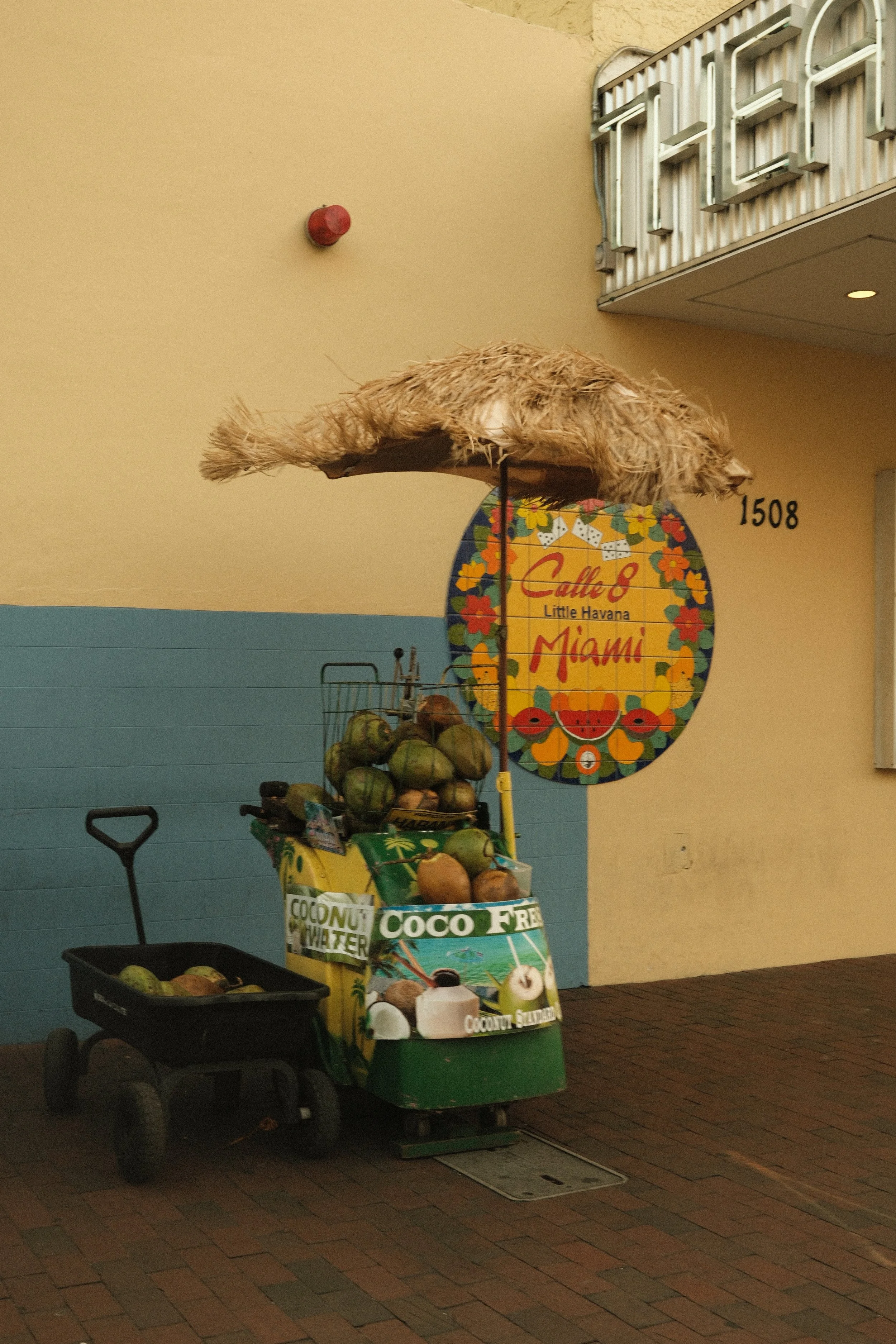 A street vendor cart selling coconuts and watermelons with a tropical design and a straw umbrella, parked near a wall with a colorful Miami-themed sign reading 'Calle 8 Little Havana Miami'.