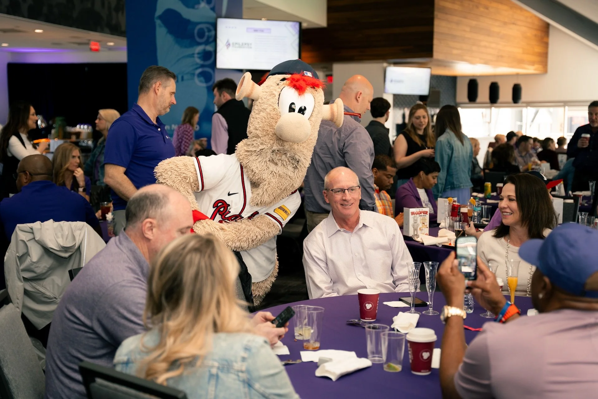 A group of people sitting at a round table in a restaurant or event space, with a mascot dressed as a bear in a baseball jersey standing and engaging with the guests. The mascot has curly fur, a baseball cap, and is smiling. Some guests are taking ph
