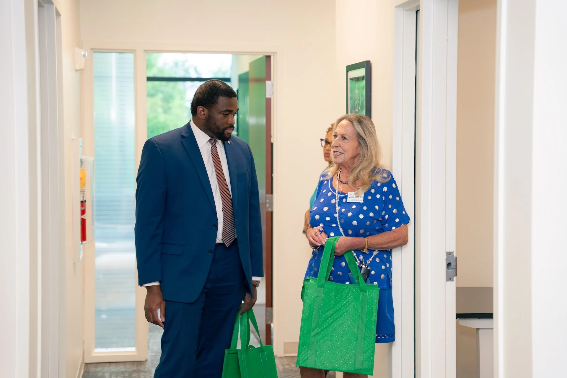 A Black man and a White woman are standing in a hallway, talking. The woman is wearing a blue polka dot dress and holding a green tote bag. The man is in a blue suit and also holding a green tote bag. Another woman is partially visible behind them.