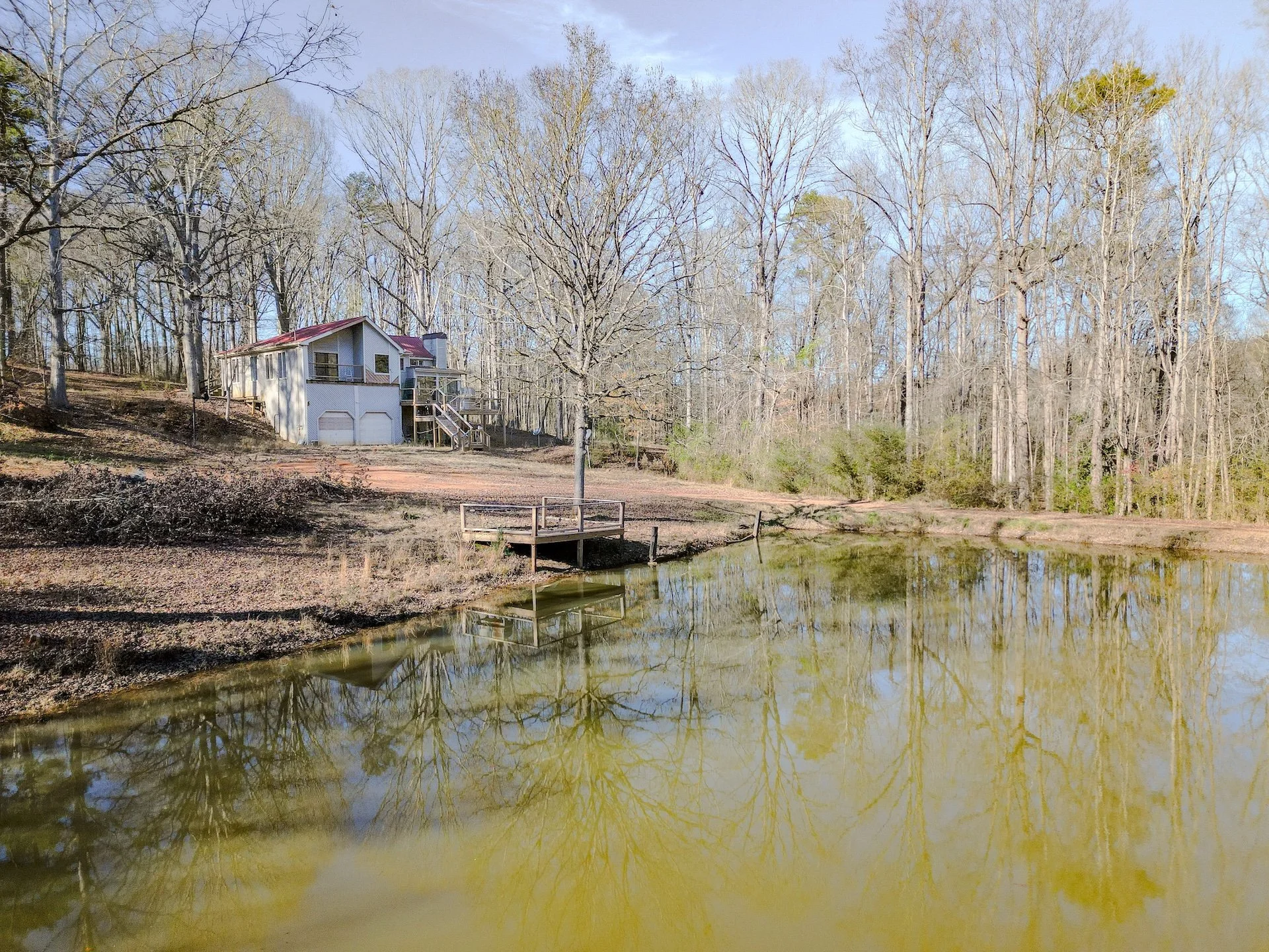 A small house with a red roof and a deck is situated on a hill near a pond, with trees that have no leaves surrounding the area.