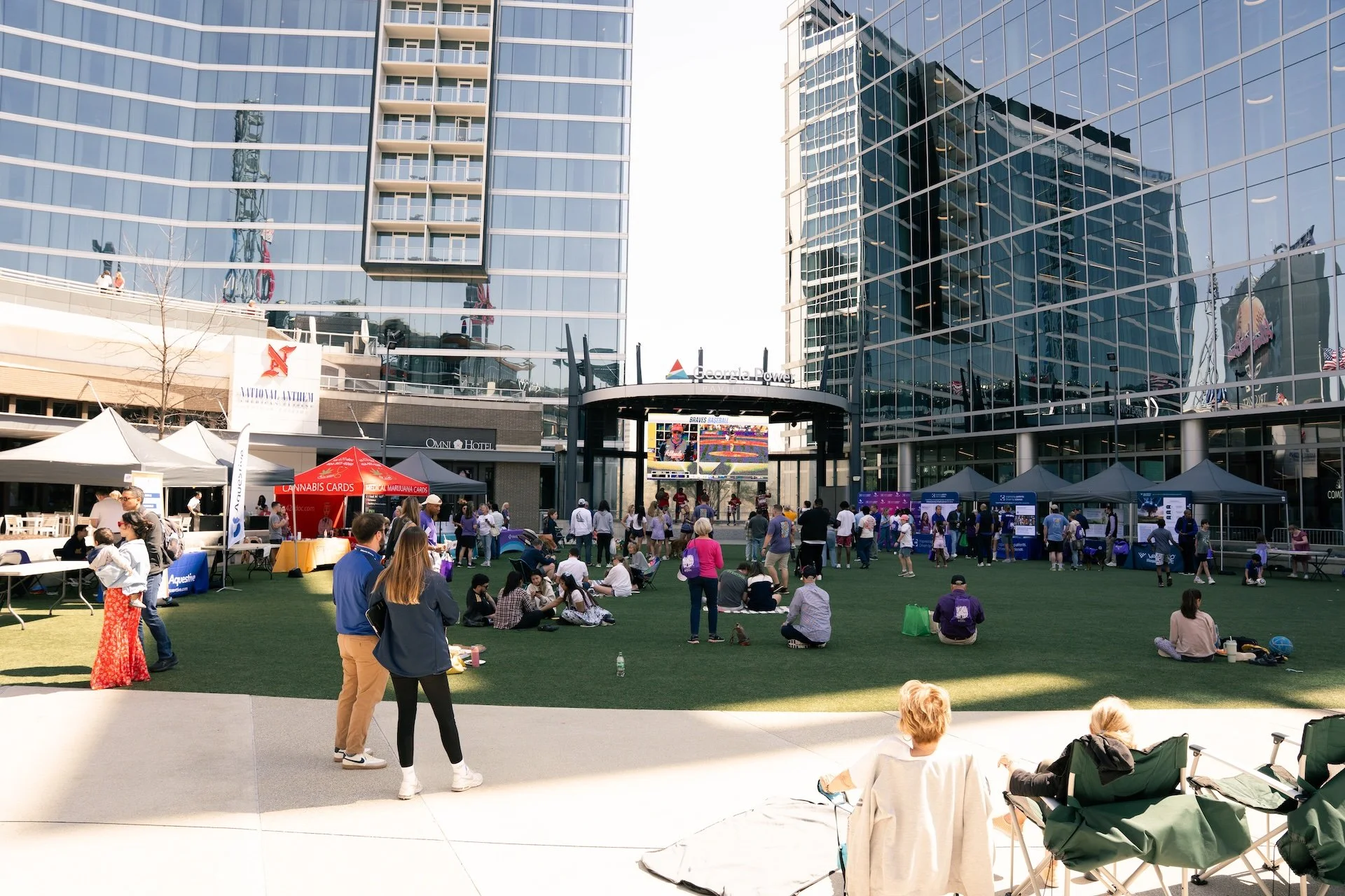 Outdoor event in a city with tents and a crowd, skyscrapers with glass windows surrounding the area, and people sitting and standing on a grassy space.