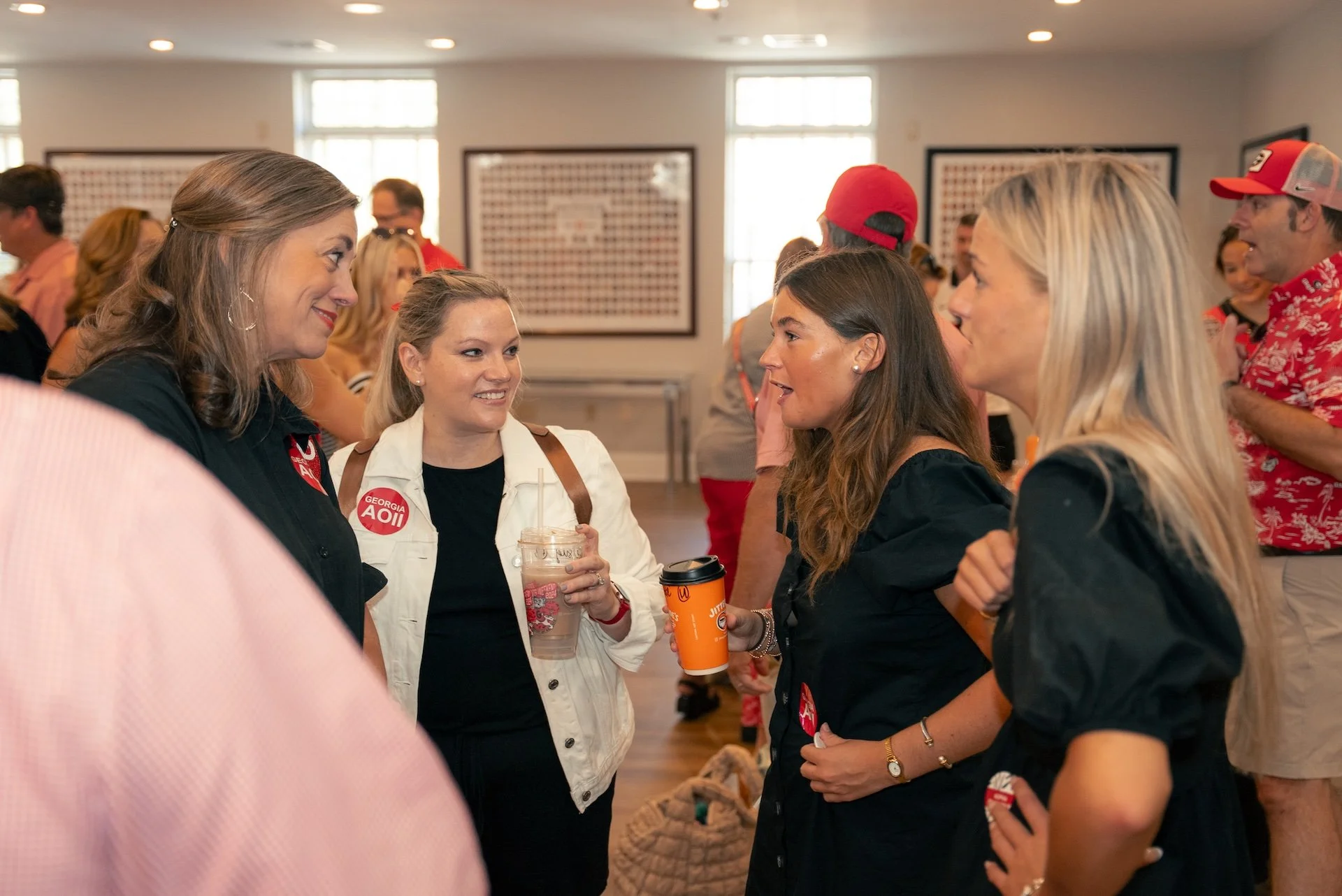 Four women engaged in conversation at an indoor event, with people in the background wearing casual and festive clothing.