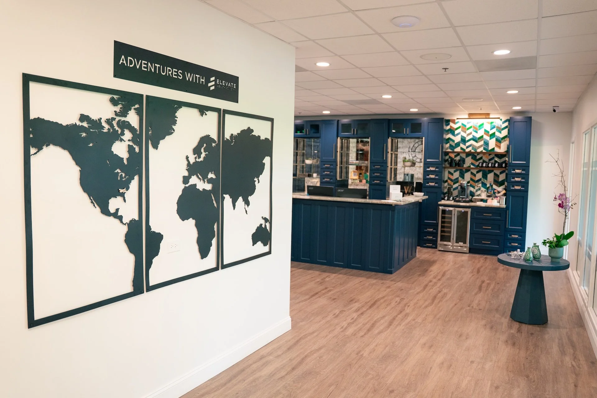 Hotel lobby with a world map wall decor, dark blue reception desk, colorful patterned wall behind the coffee station, and a small table with decorative items and a potted orchid.
