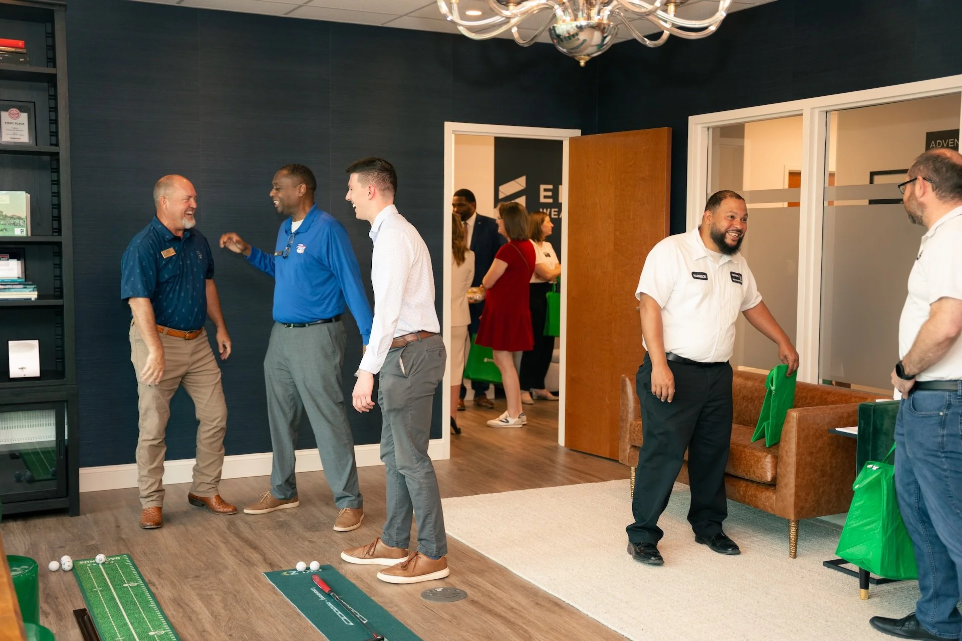 People socializing in an office space with golf putting mats on the floor, including men and women smiling, talking, and laughing. The room has dark blue walls, bookshelf, a chandelier, and a brown couch with green gift bags.