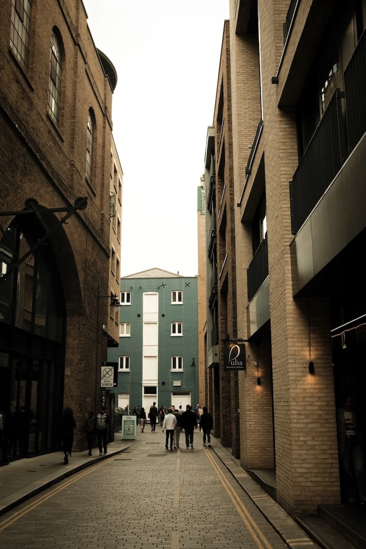 Pedestrians walking down a narrow city alleyway lined with brick buildings on both sides, with a tall modern building at the end of the alley.