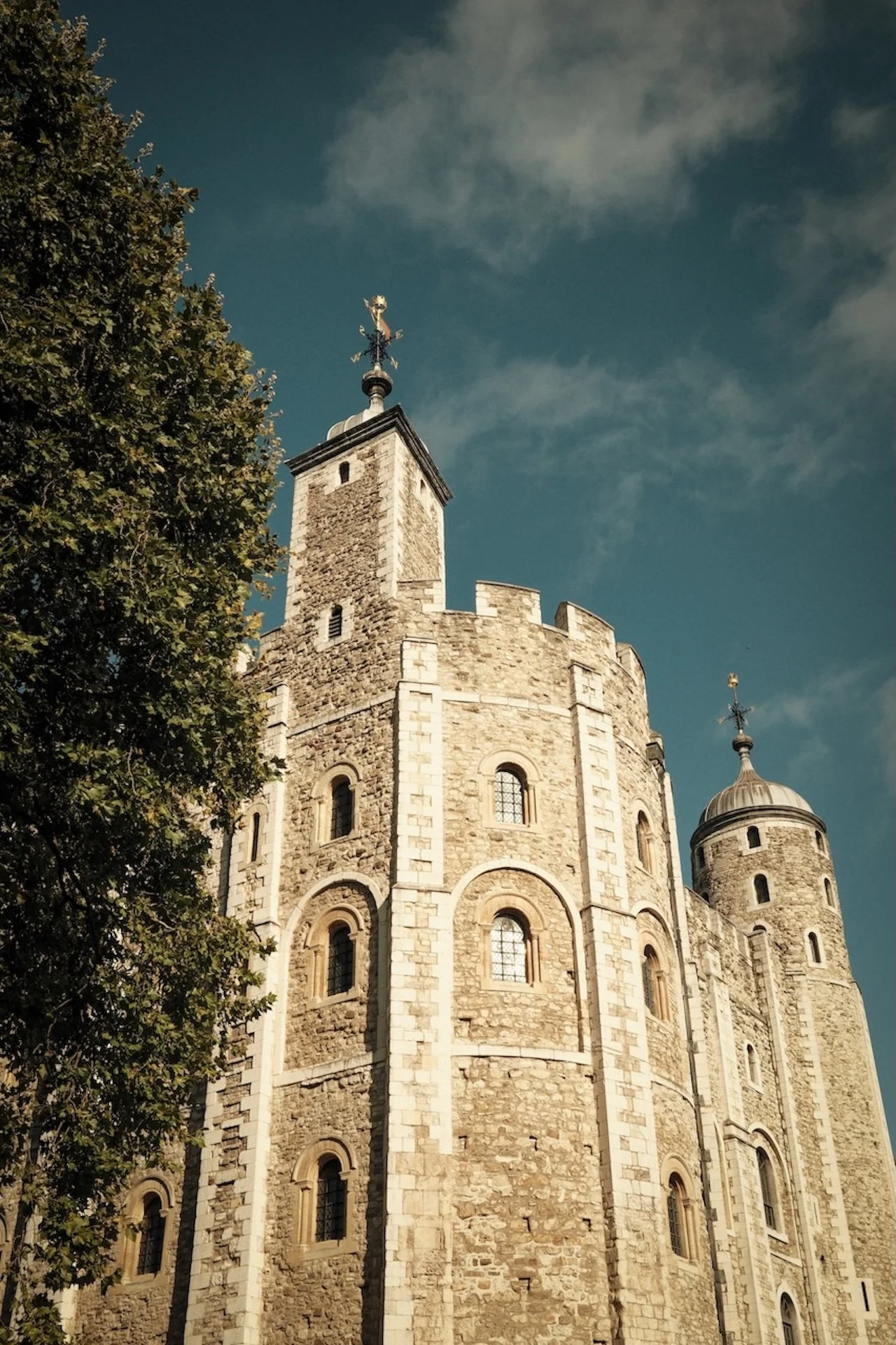 A historic stone castle with multiple towers and small windows, topped with weather vanes and crosses, under a partly cloudy blue sky, with a tree on the left side.