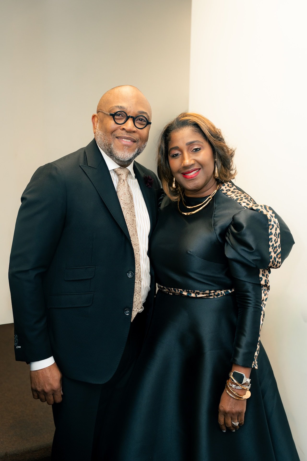 A man and woman posing together, dressed in formal black attire. The man is wearing glasses, a suit with a tie, and has a beard. The woman is wearing jewelry and a dress with leopard print accents on the sleeves and waist.
