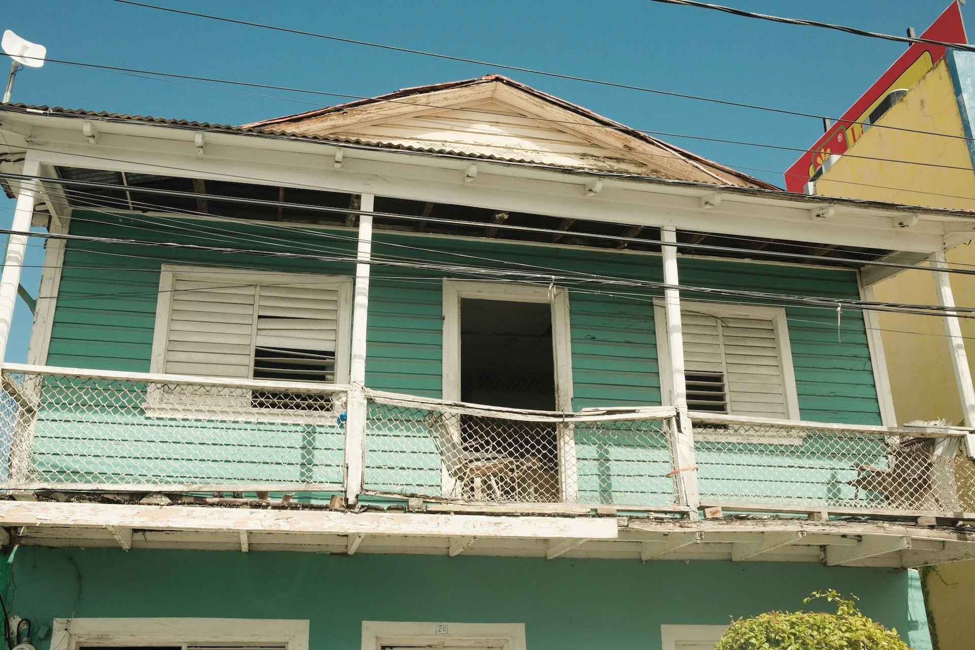 A two-story house with a teal exterior, white trim, and a weathered balcony with a metal railing. The windows have white shutters, one of which is open. The roof is slightly damaged, exposing wood underneath. The house is in a sunny area with a clear