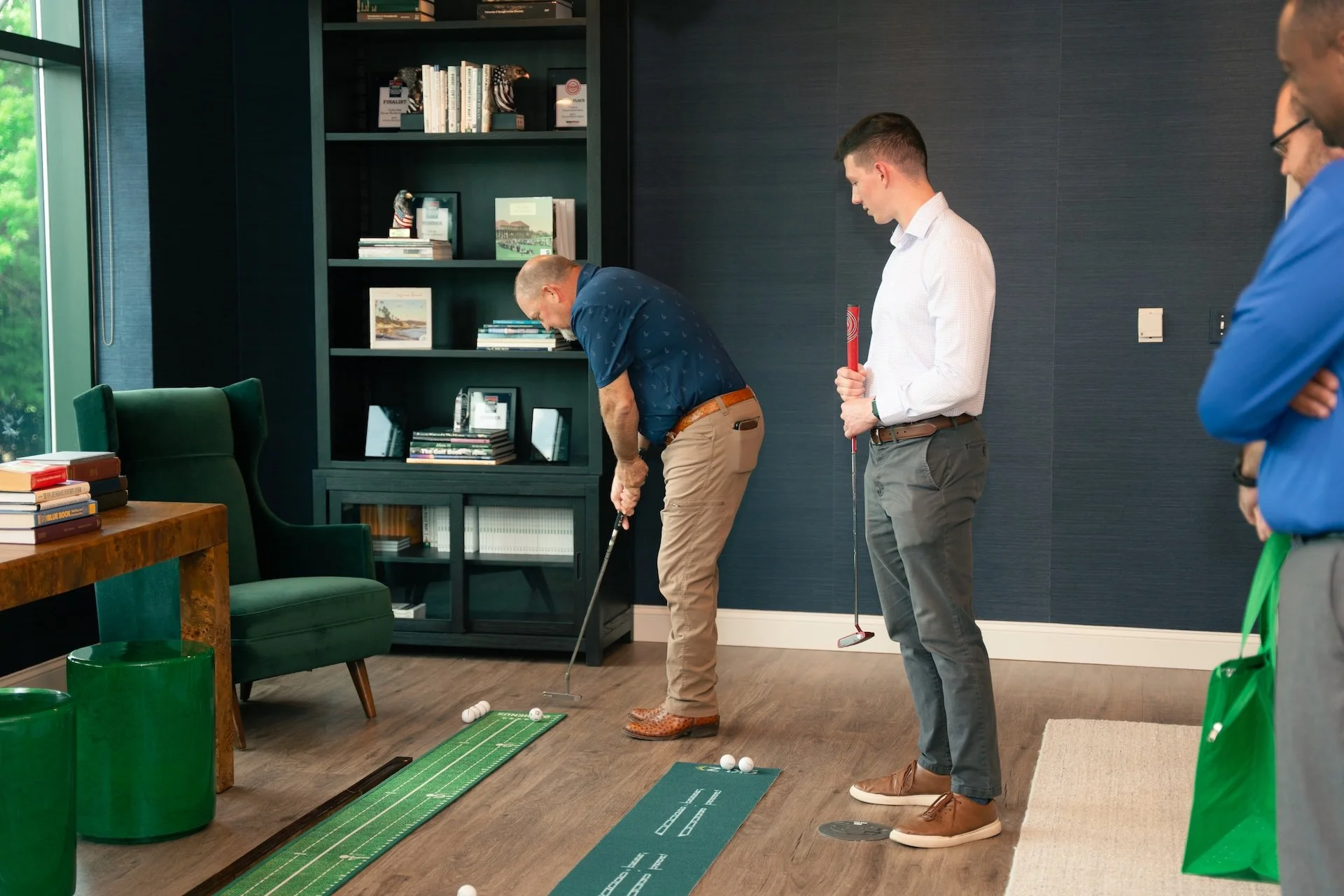 People practicing golf indoors, with one person taking a shot and others observing, using miniature golf putting mats.