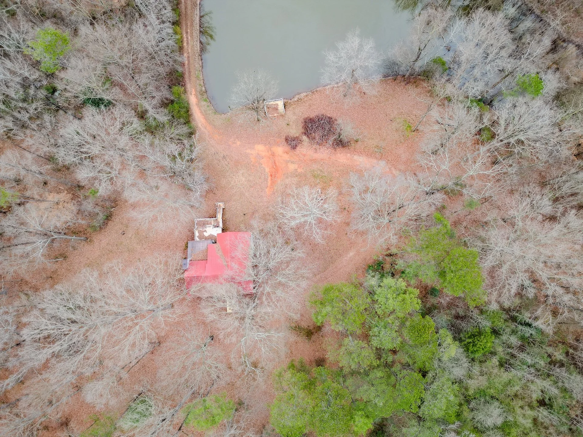 Aerial view of a red-roofed house surrounded by leafless trees, a small pond, and a dirt path in a wooded area.