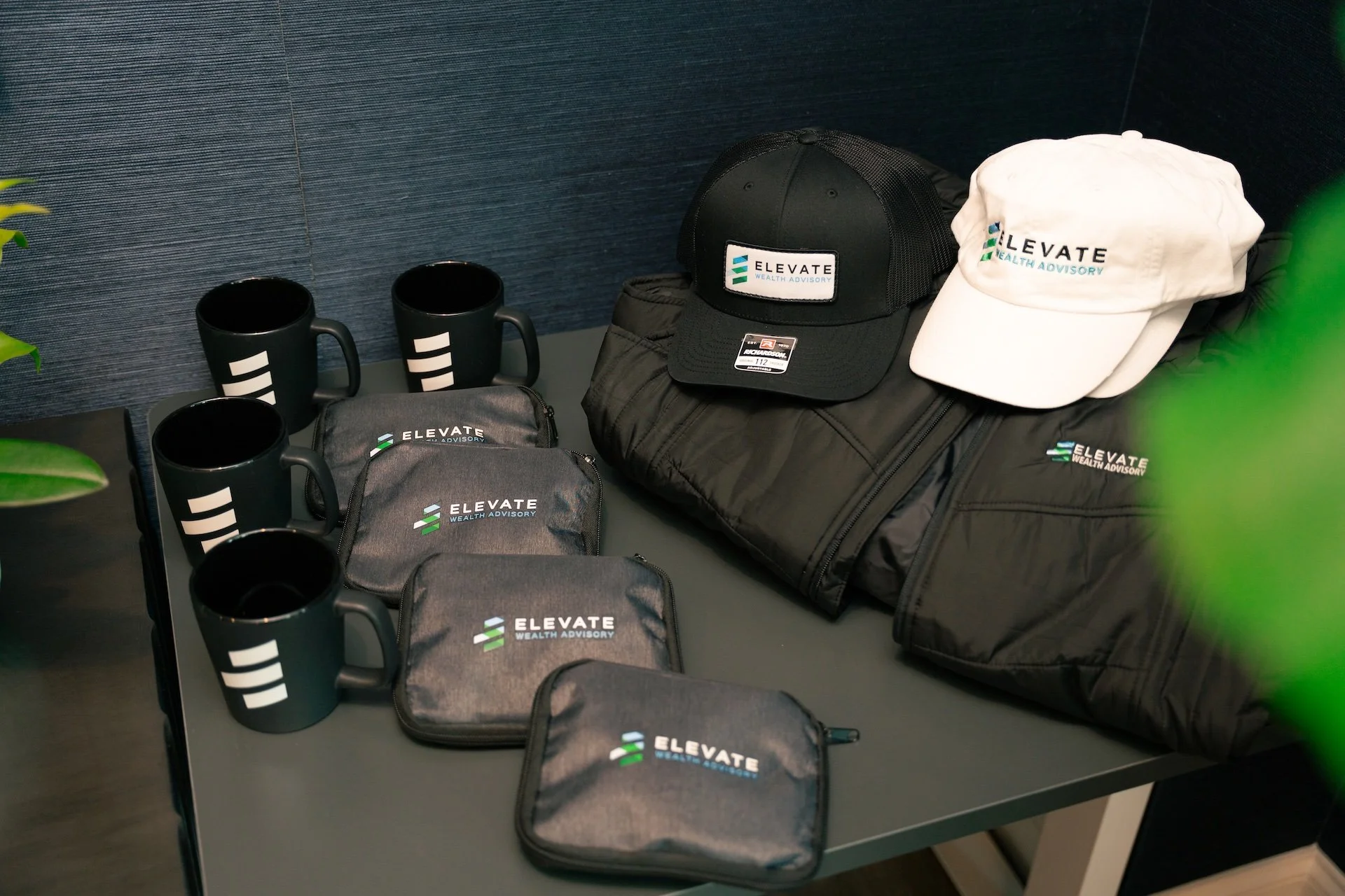 Branded promotional items including black and white hats, black jackets, black mugs, and grey pouches with the 'Elevate Wealth Advisory' logo displayed on a table.
