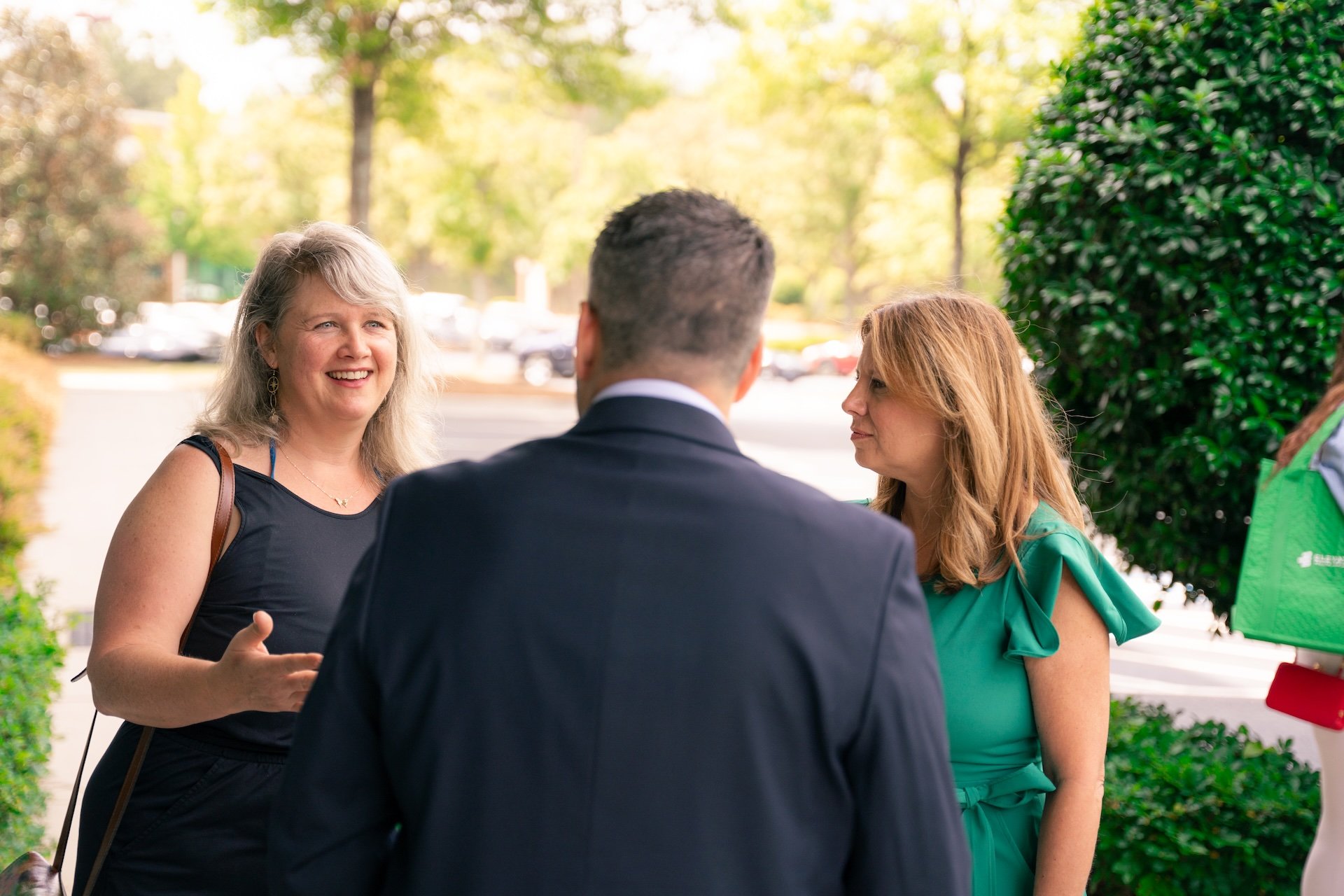 Three people talking outdoors near a parking lot and green bushes, two women and one man, smiling and engaged in conversation.