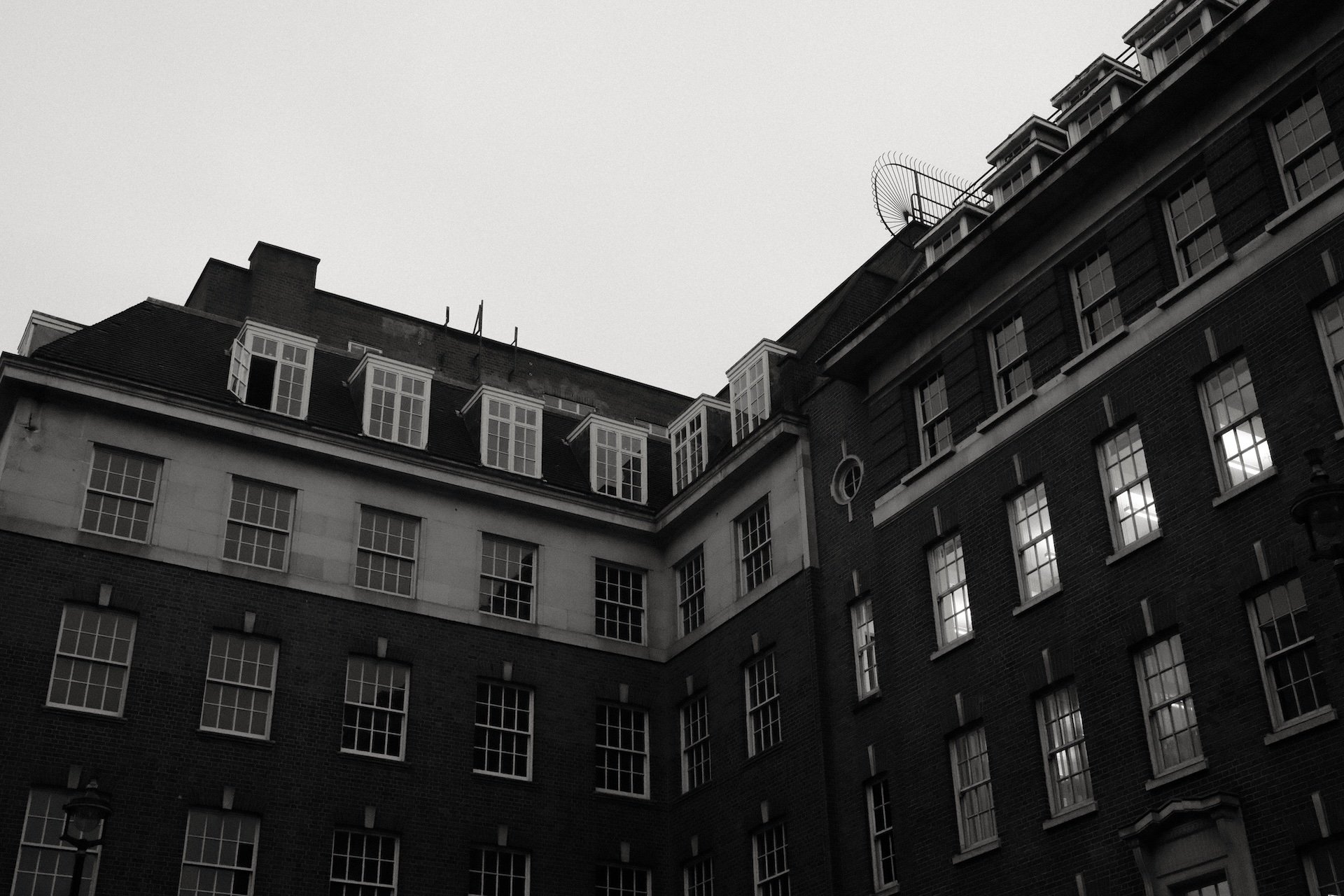 Black and white photo of multi-story brick and stone buildings with many windows, some with lights on, under a cloudy sky.