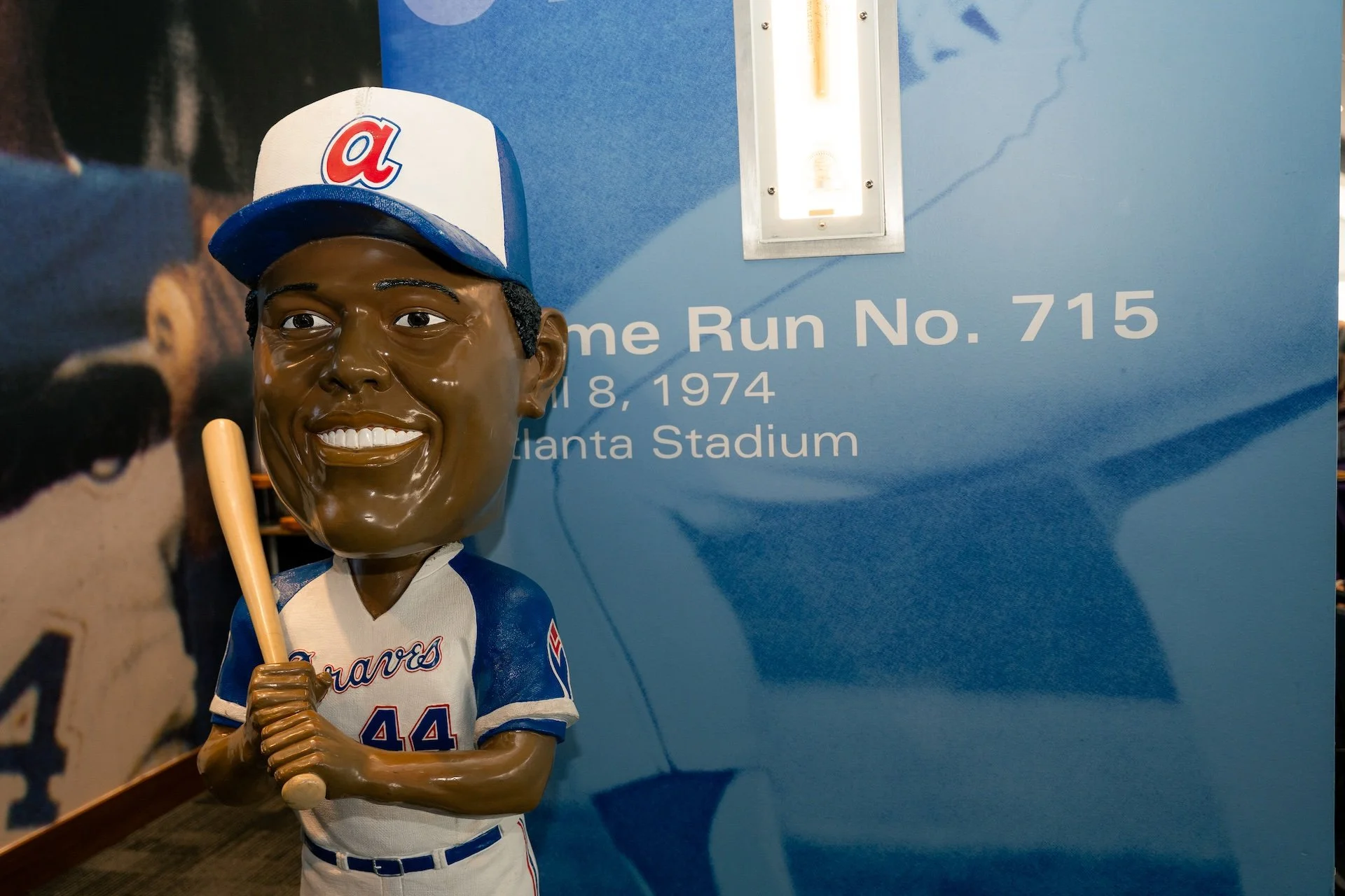 A bobblehead figurine of a baseball player in a Braves uniform, wearing a baseball cap, holding a bat, and smiling, displayed in front of a blue informational background about Hank Aaron's 715th home run at Atlanta Stadium.