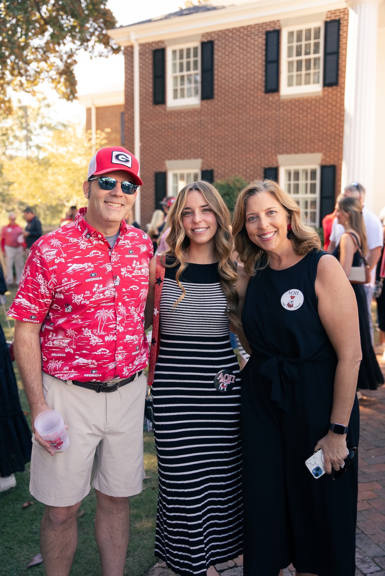 A group of three people, two women and a man, standing outdoors at a graduation event. They are smiling and dressed in casual summer clothes. The man on the left is wearing a red baseball cap, sunglasses, a red Hawaiian shirt with a Georgia Bulldogs 