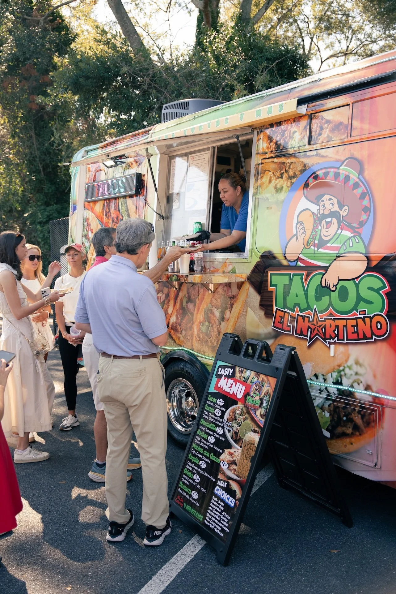 People ordering food at a taco food truck named 'Tacos El Norteño' during daytime, with a woman serving from the window. A menu board is displayed outside, and there are trees in the background.
