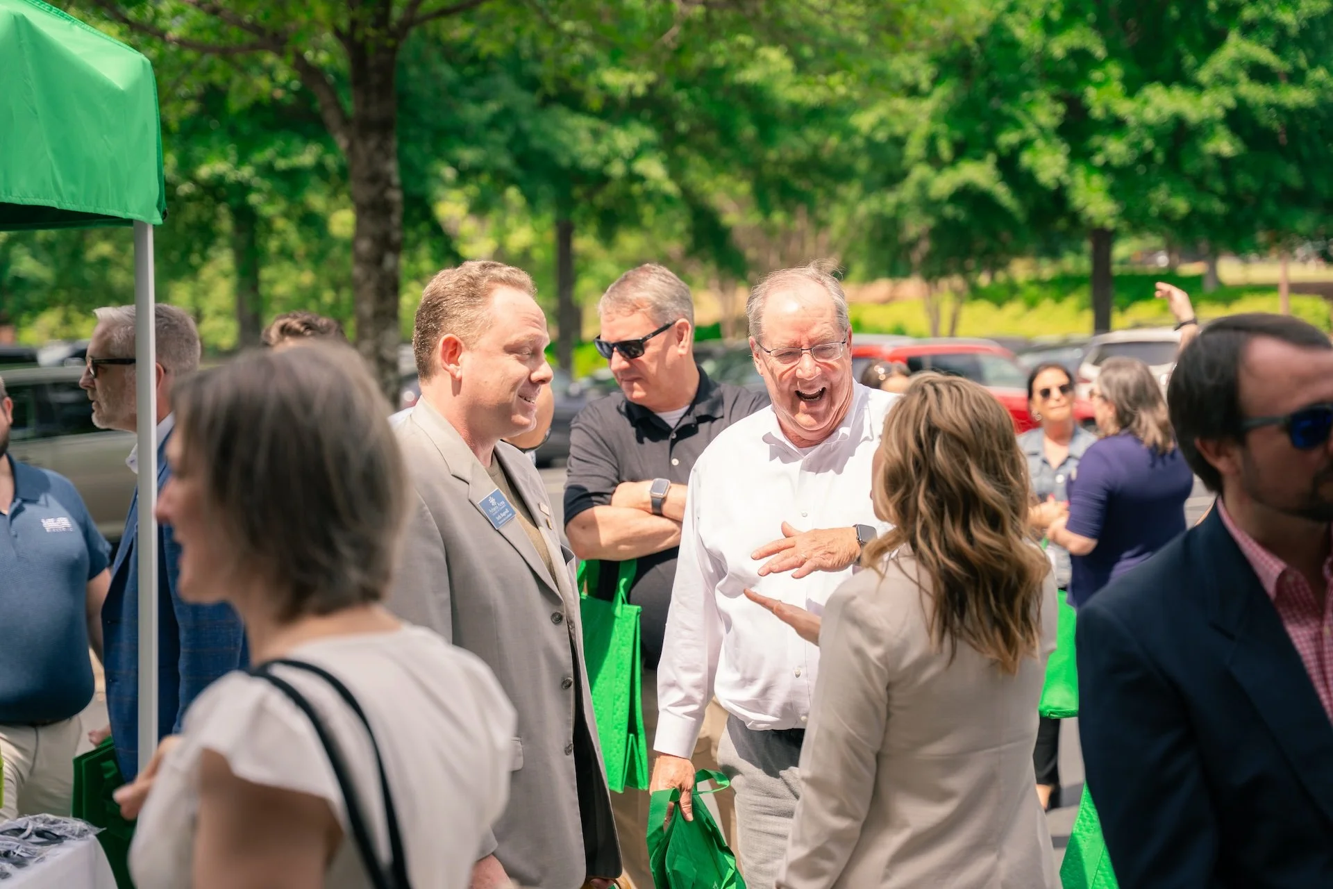 People engaging in a lively conversation outdoors at an event, with trees and parked cars in the background.
