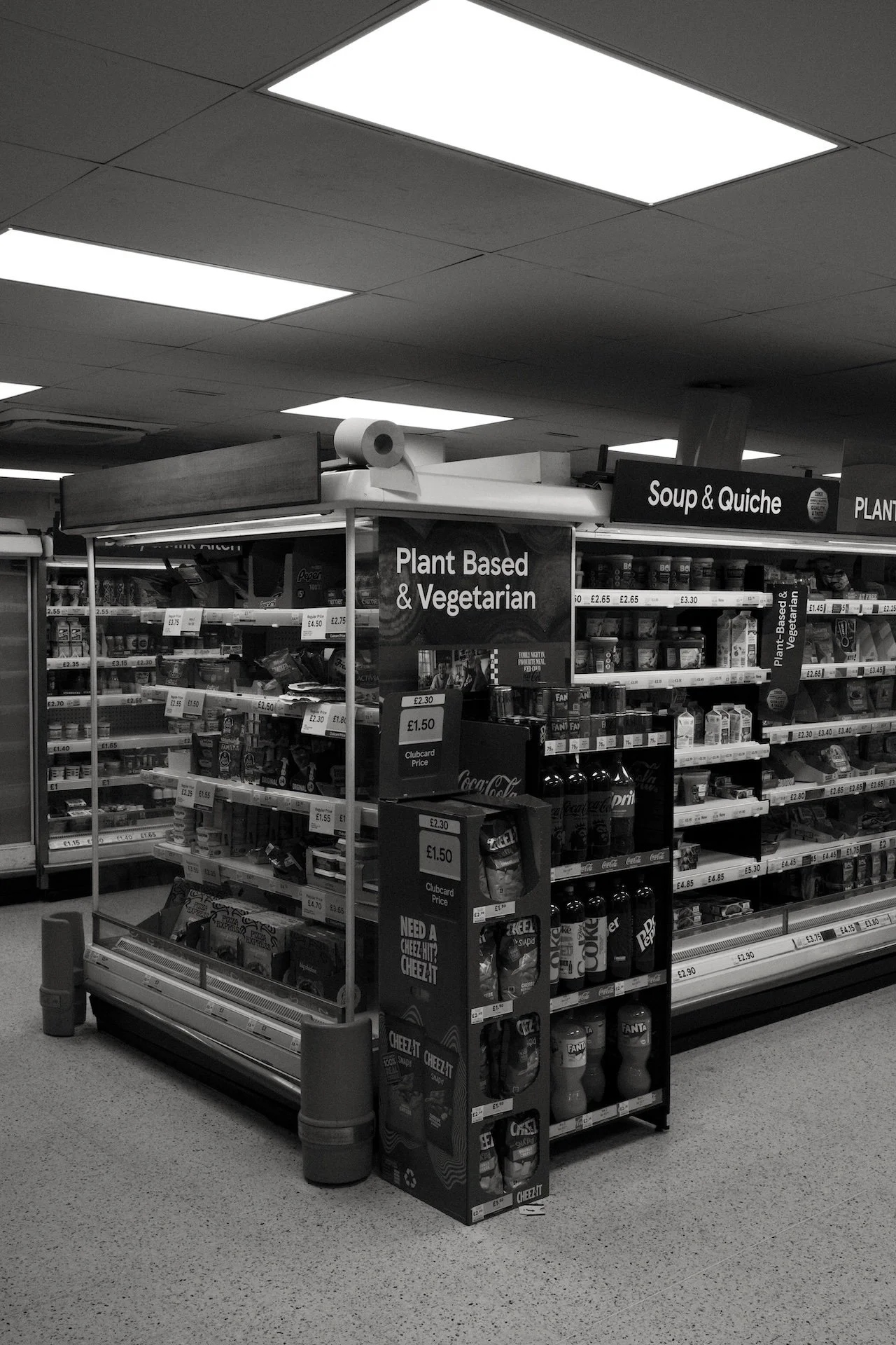 A supermarket aisle section labeled 'Plant Based & Vegetarian' and 'Soup & Quiche,' displaying shelves with various food products, soda bottles, and snack items. The area has overhead fluorescent lighting.