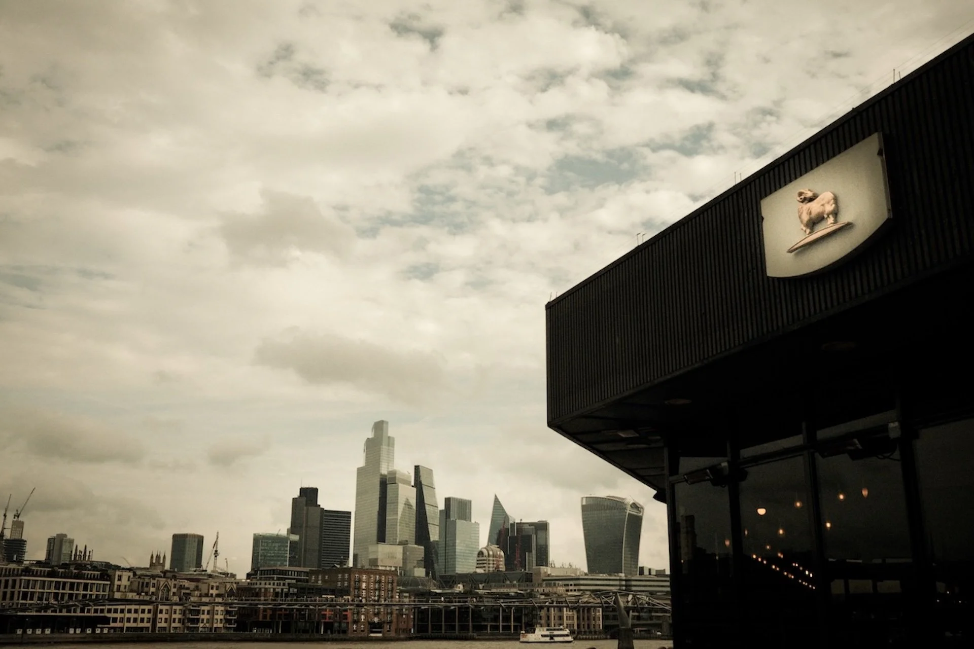 City skyline with tall buildings, some under construction, on a cloudy day. Part of a modern building with a dog statue on a sign is in the foreground on the right.