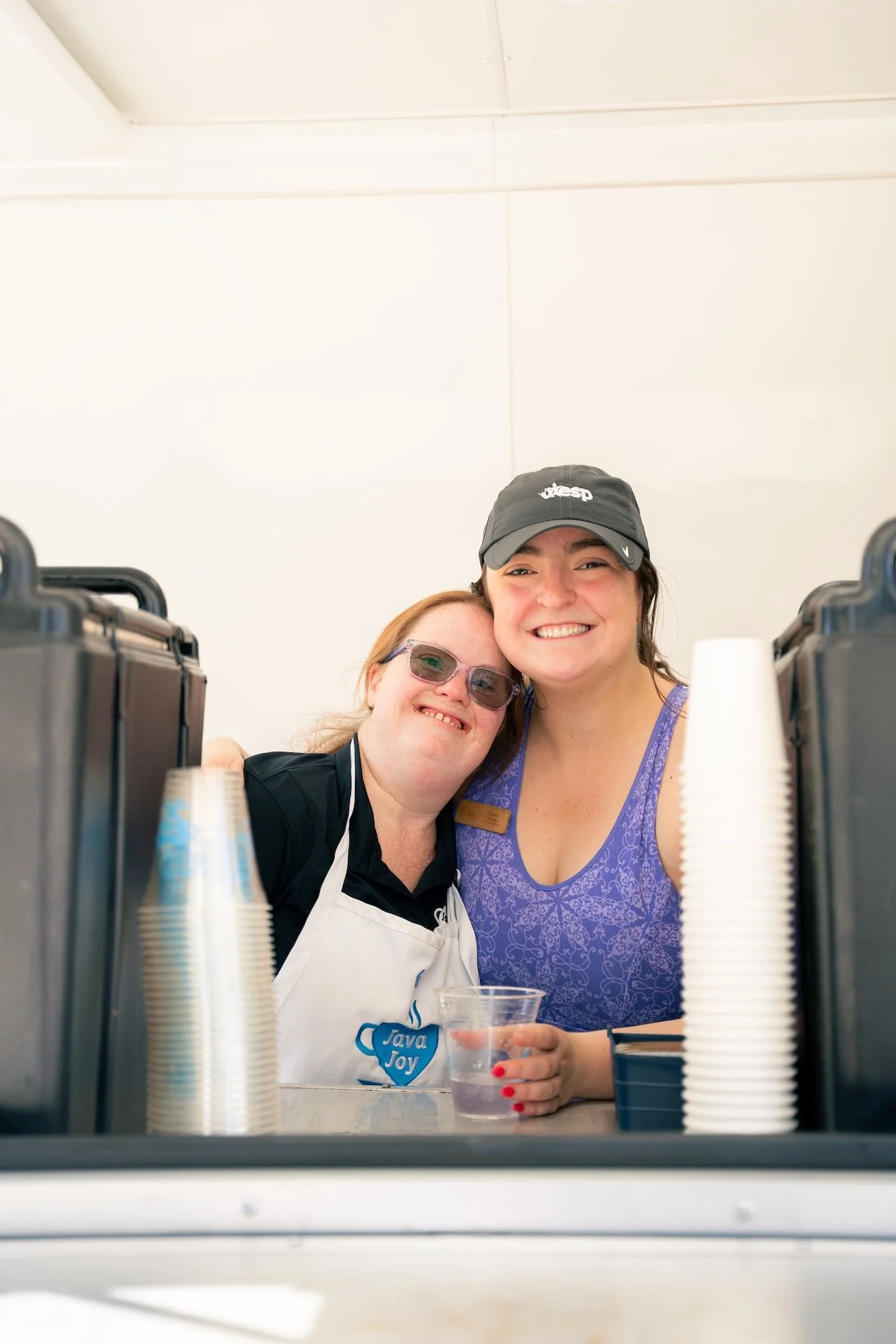 Two women smiling and hugging at a beverage stand, with disposable cups and water in a clear cup in front of them.