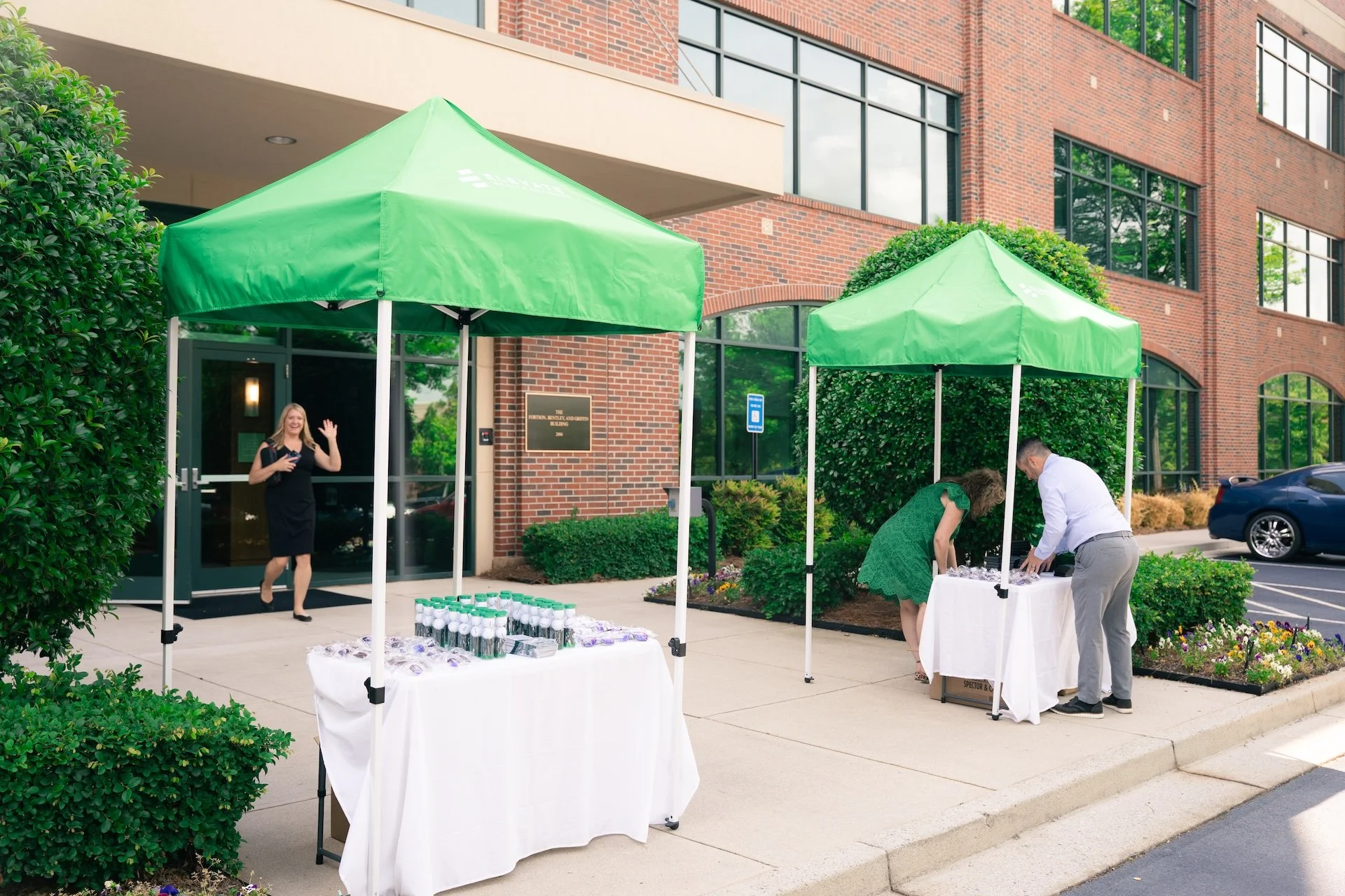 People setting up a table for an event outside a brick building with green tents, supplies, and flowers.