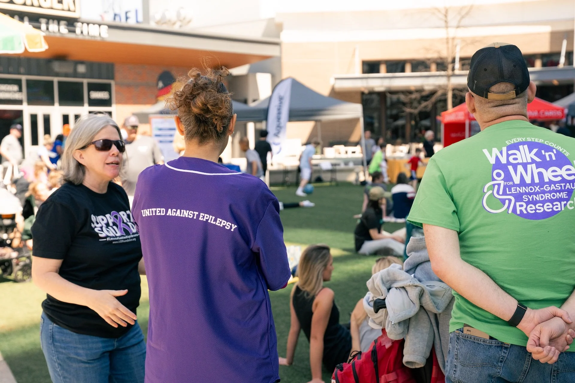 People gathered outdoors for a walk-and-wheel event supporting research for Lennox-Gastaut syndrome, with participants and spectators by a building and tents.