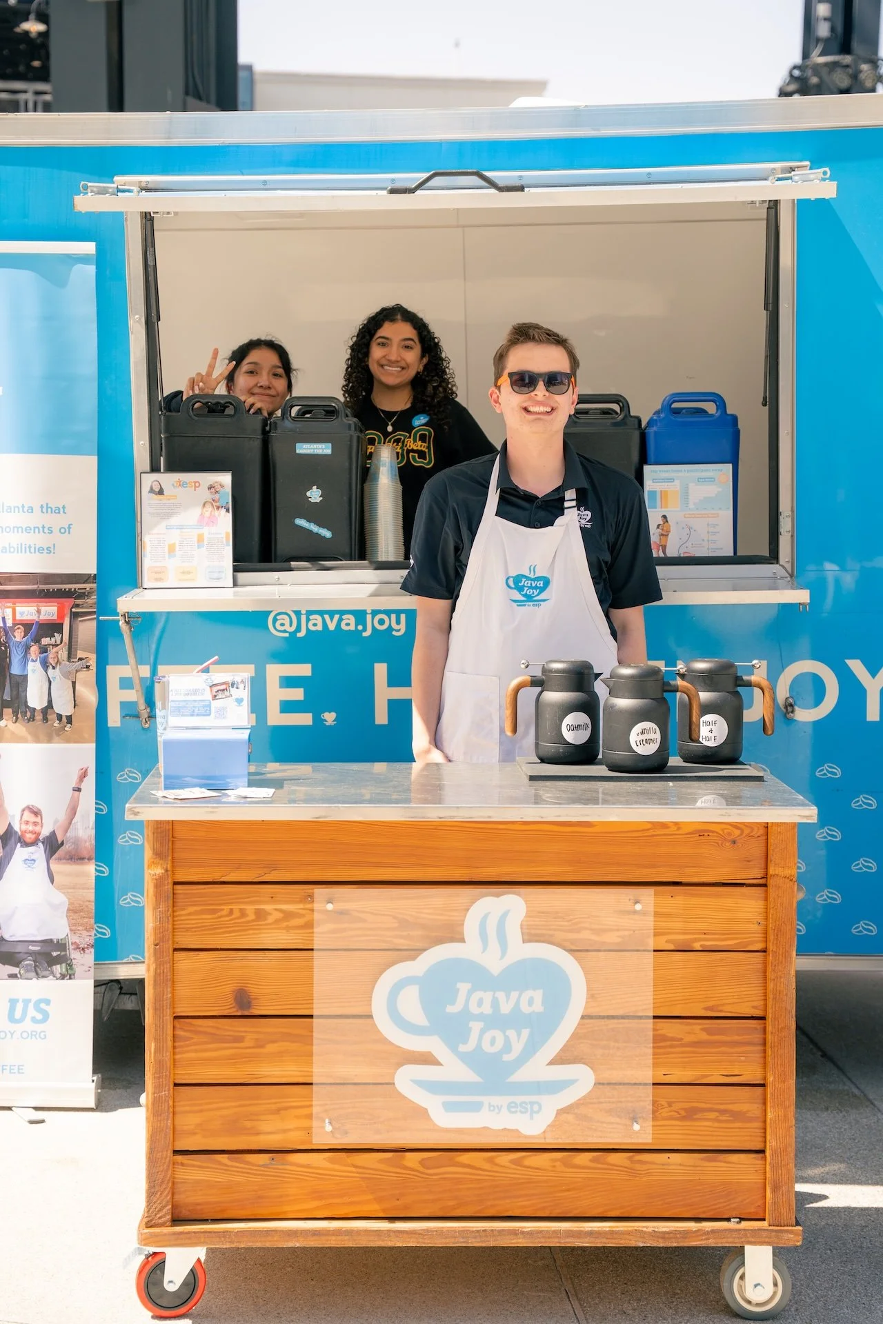 A young man with sunglasses and a white apron standing in front of a blue mobile coffee stand with three black coffee kettles labeled 'Oat Milk,' 'Vanilla or Caramel,' and 'Half & Half.' Behind him are two smiling women inside the stand.