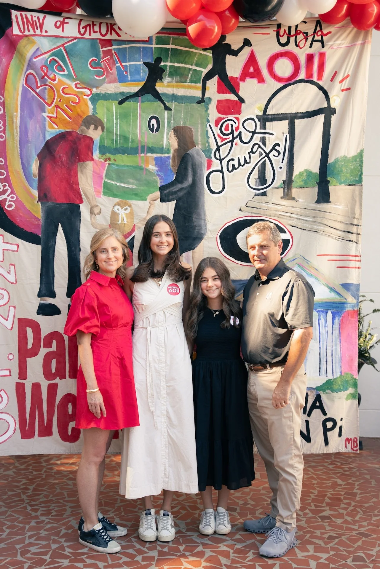 Four people standing in front of a colorful, artistic backdrop with school-related drawings and words such as "Go Dawgs!" and "Be Miss S". The group includes one older woman, two teenage girls, and one adult man. The backdrop features balloons at the
