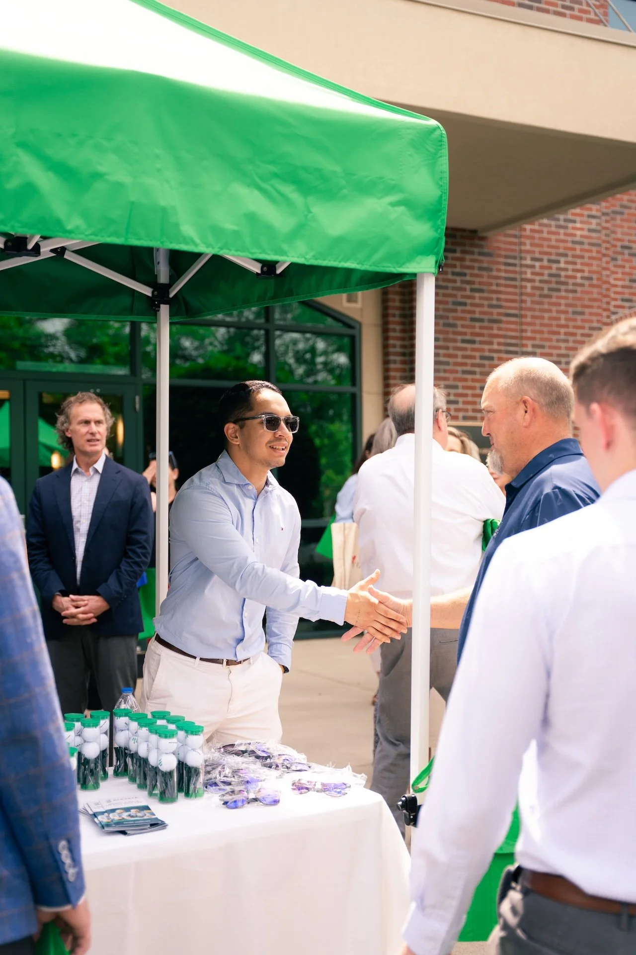 Man in sunglasses shaking hands at a table with golf-related items during an outdoor event.