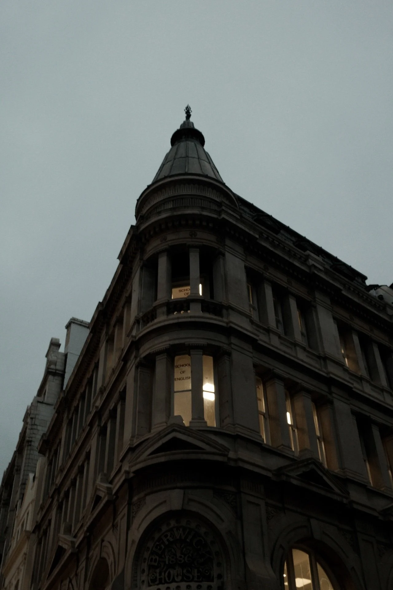A tall, historic stone building with a rounded corner and a conical tower on top, illuminated windows, and a cloudy sky in the background.