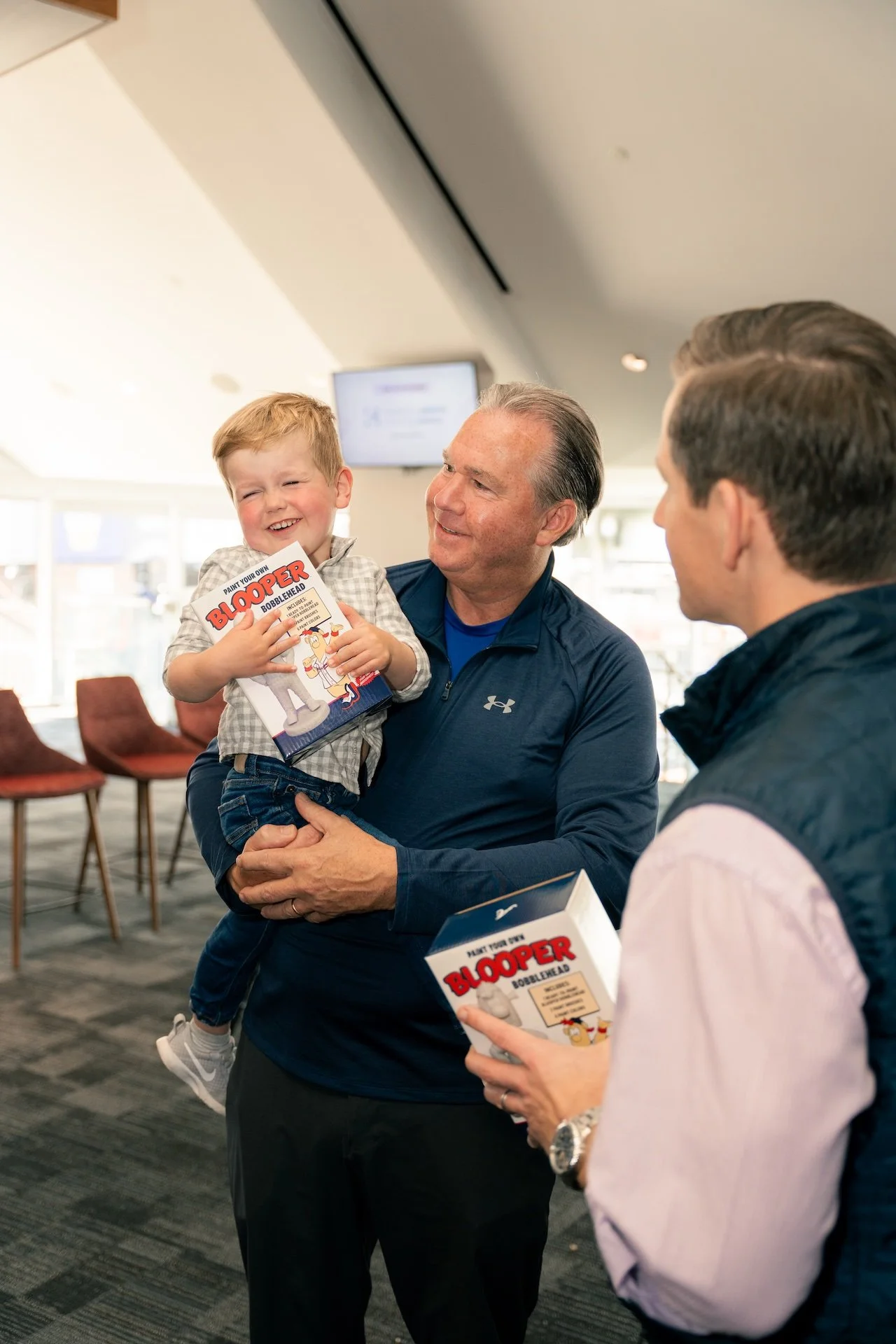 A young boy being held by an older man, both smiling and holding a game box titled 'Bloopers Bobblehead'. Another man is standing nearby, also holding a similar game box, in a brightly lit indoor space with brown chairs in the background.