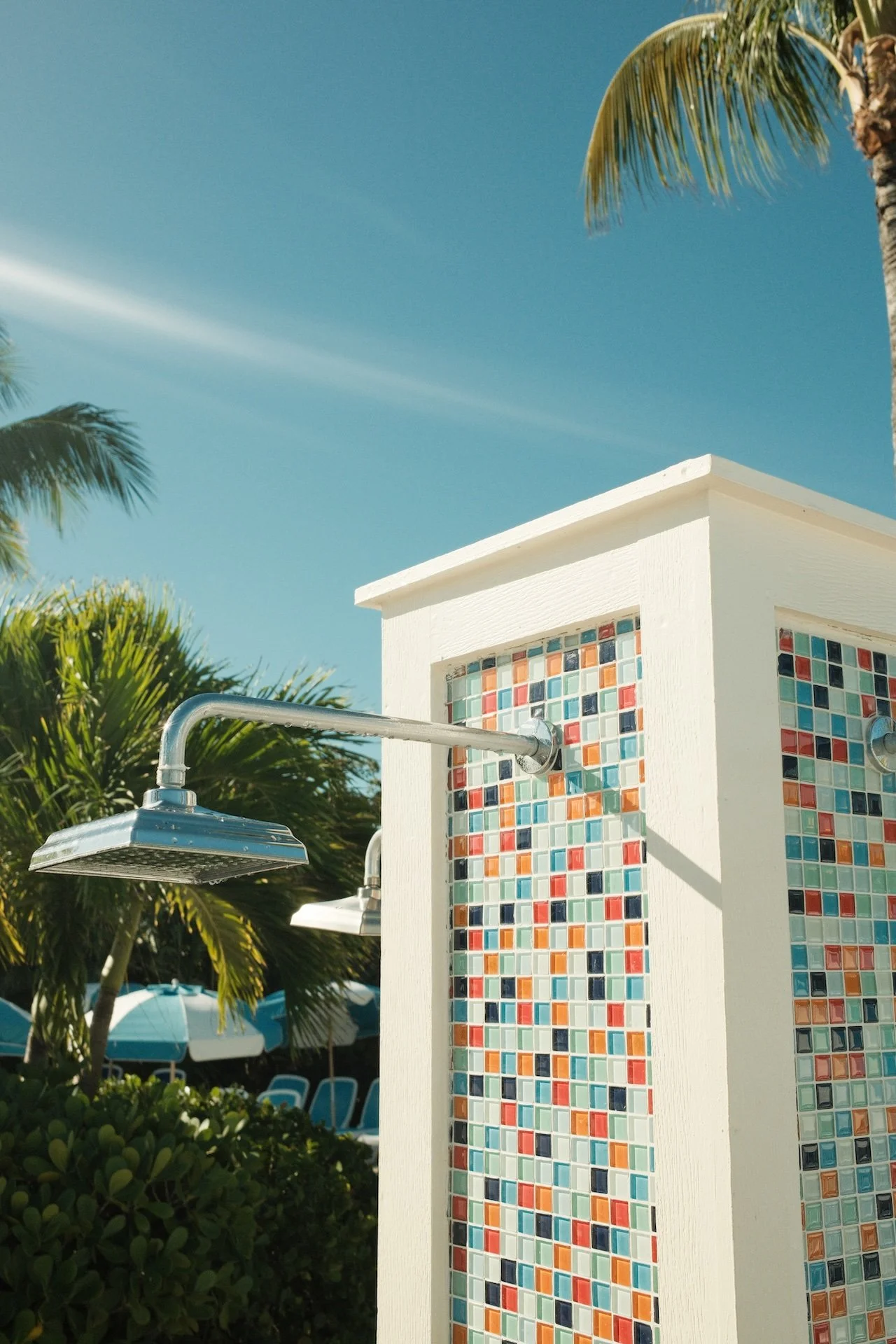 Outdoor shower with colorful mosaic tiles, palm trees, and blue sky in the background.